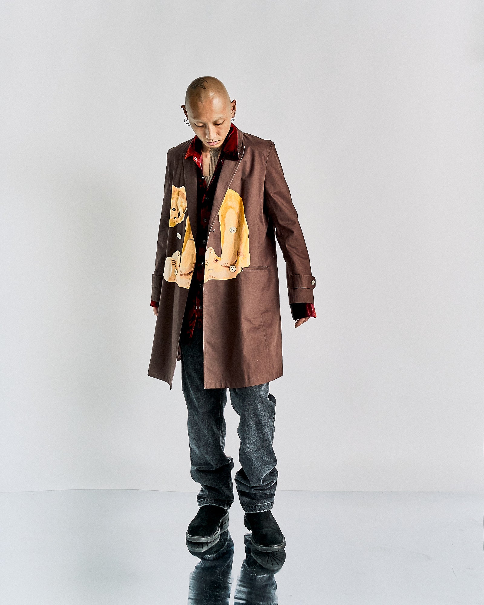 Person stands in a brown John Undercover coat with cartoon imagery, looking down, set against a white background. They wear dark jeans and black shoes, exuding a reflective mood.