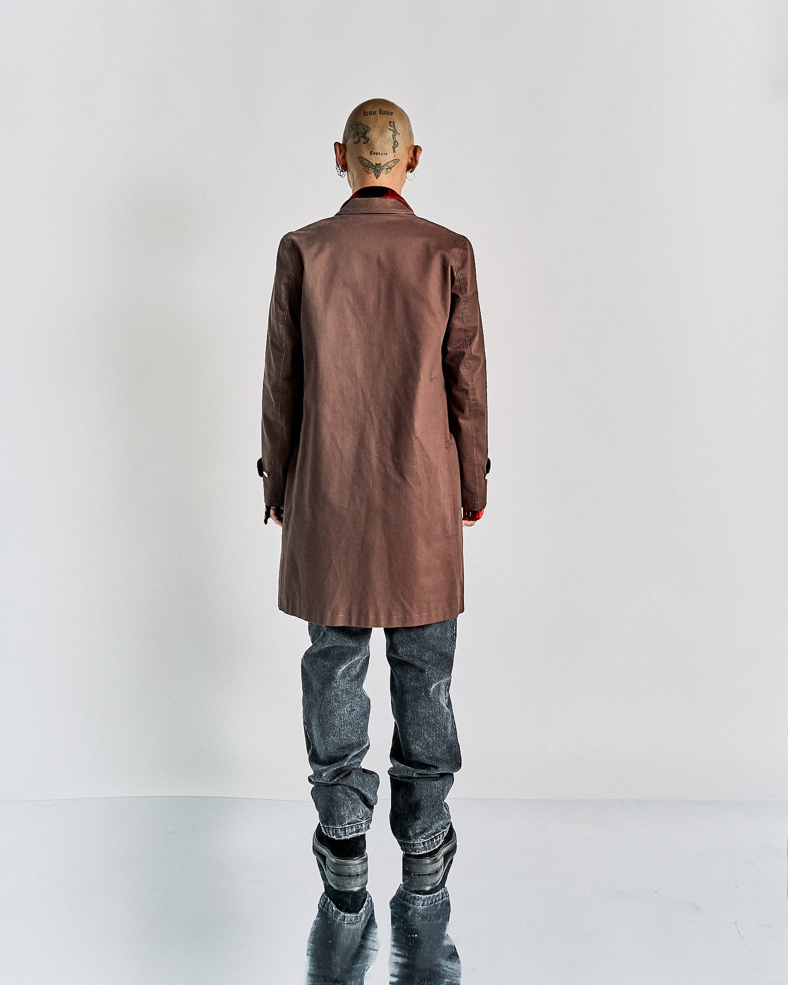 A person with a buzzed head and tattoos stands facing away, wearing a brown John Undercover coat and jeans. The backdrop is minimalist white, reflecting a modern, edgy style.