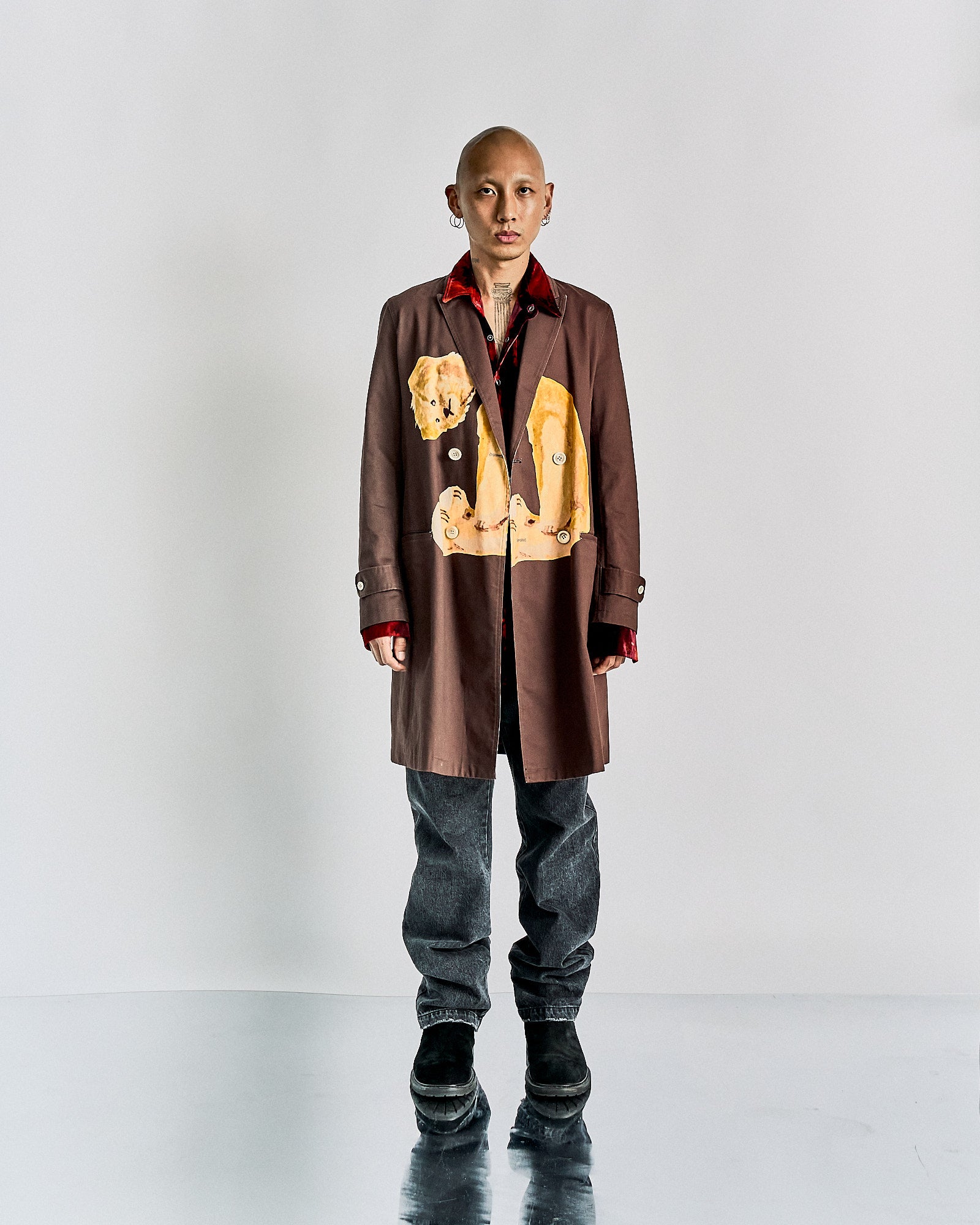 A person standing against a white background, wearing a double-breasted John Undercover chester coat with a graphic of a headless teddy bear on the front, paired with grey trousers and black boots.