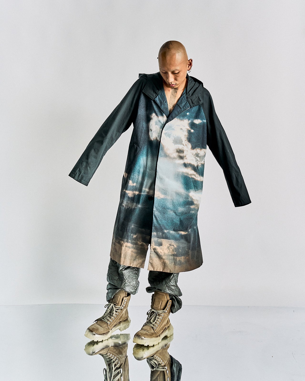 A person standing wearing a black John Undercover hooded coat with a landscape panel featuring a cloud and sky pattern. The coat has a button-up front and hip pockets.