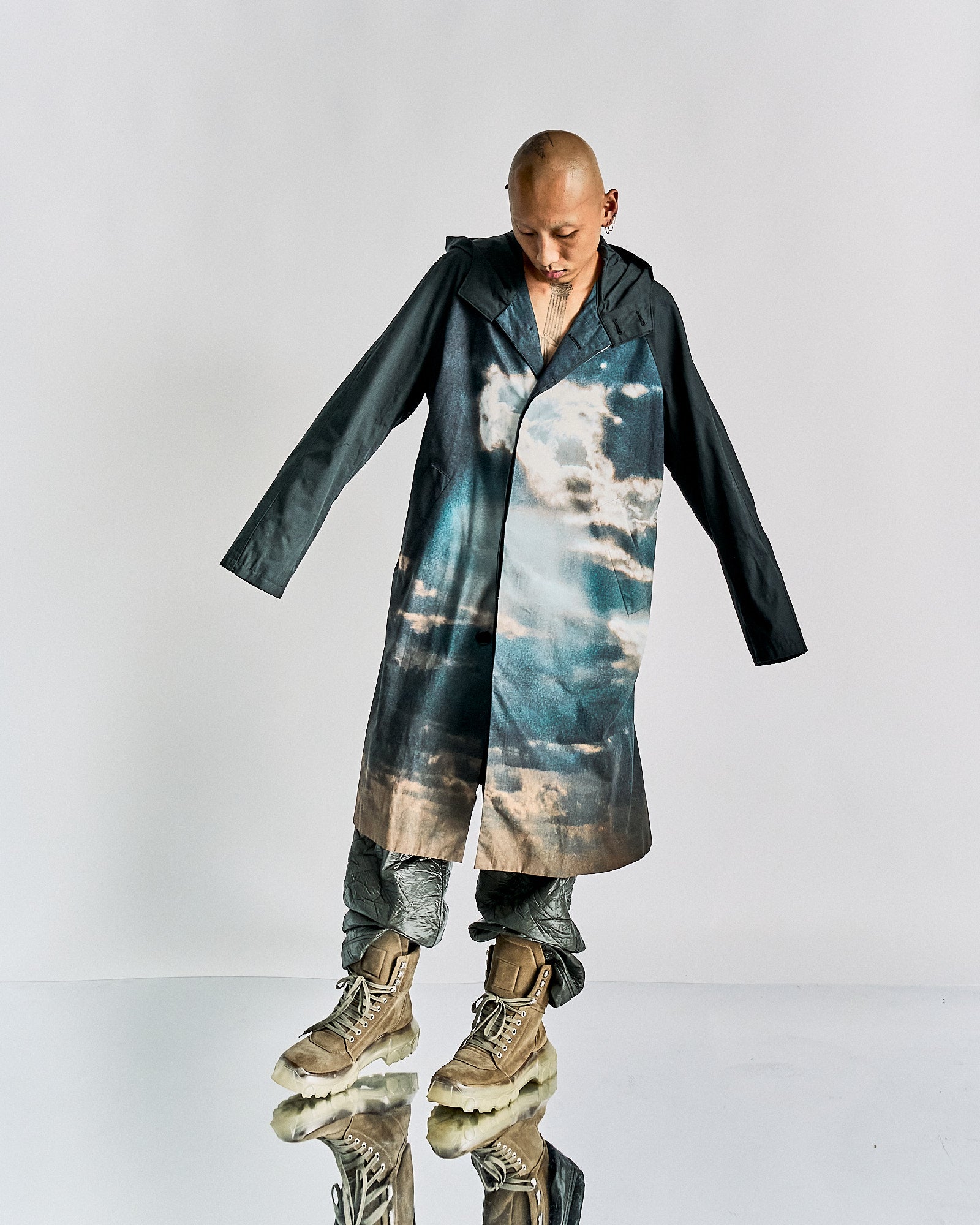 A person stands wearing a John Undercover coat with a sky pattern, pairing it with metallic pants and brown, rugged boots. They appear contemplative, looking down.