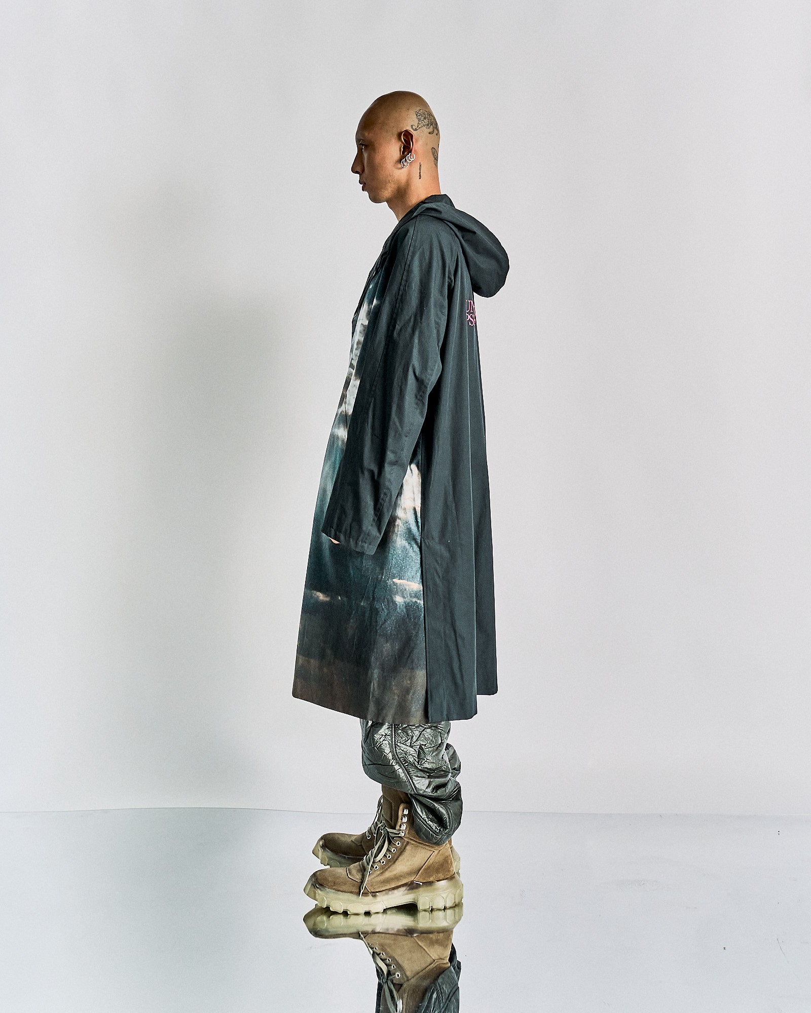 A person in profile wearing a long, dark hooded John Undercover coat with a printed design, metallic pants, and chunky, layered boots stands against a plain background. The tone is futuristic and edgy.