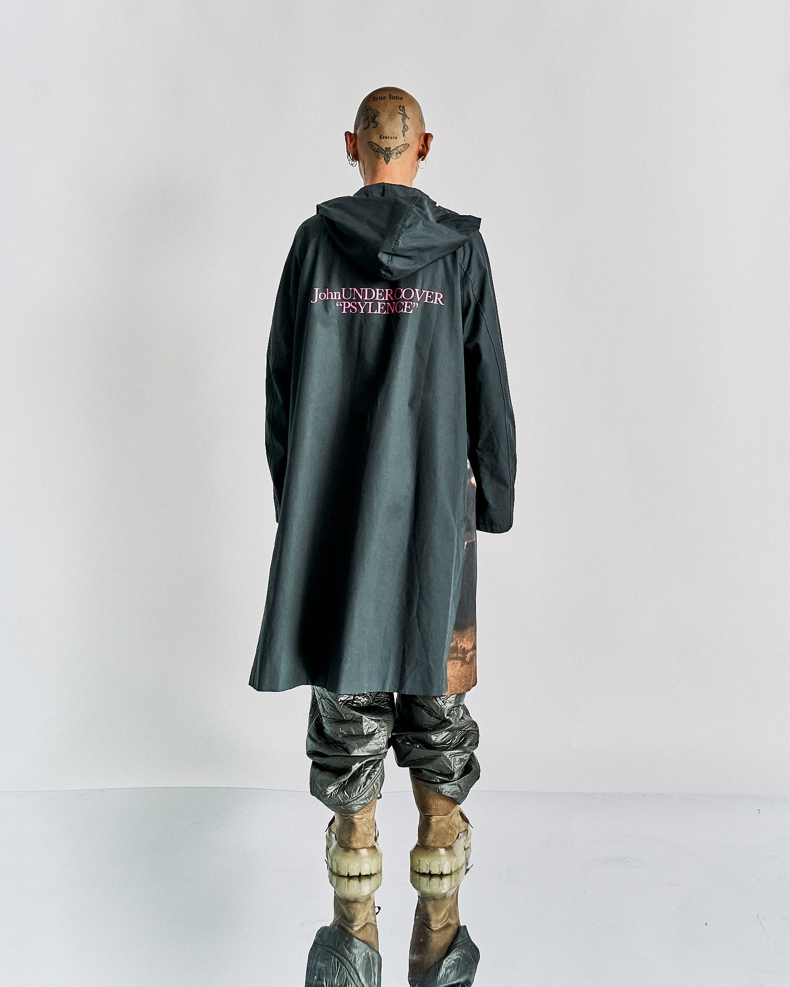 A person stands facing away, wearing a black hooded coat with "John UNDERCOVER PSYENCE" in pink on the back. They have tattooed head, metallic pants, and unique shoes.