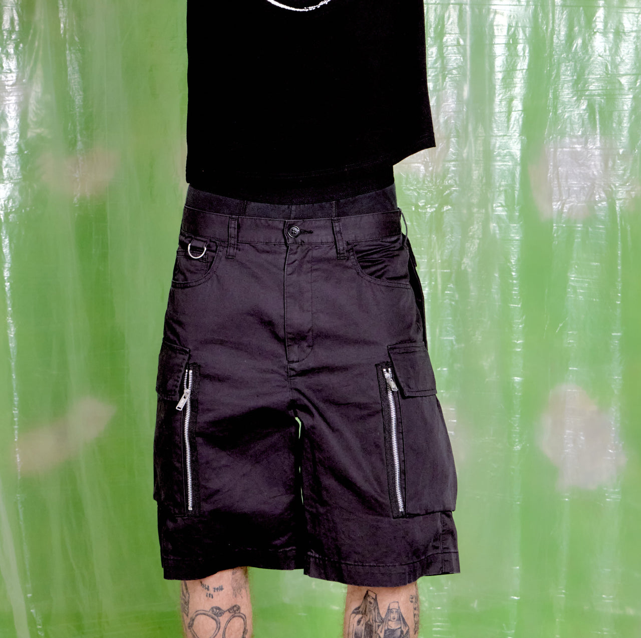 Person wearing black cargo shorts with zippers against a green curtain background