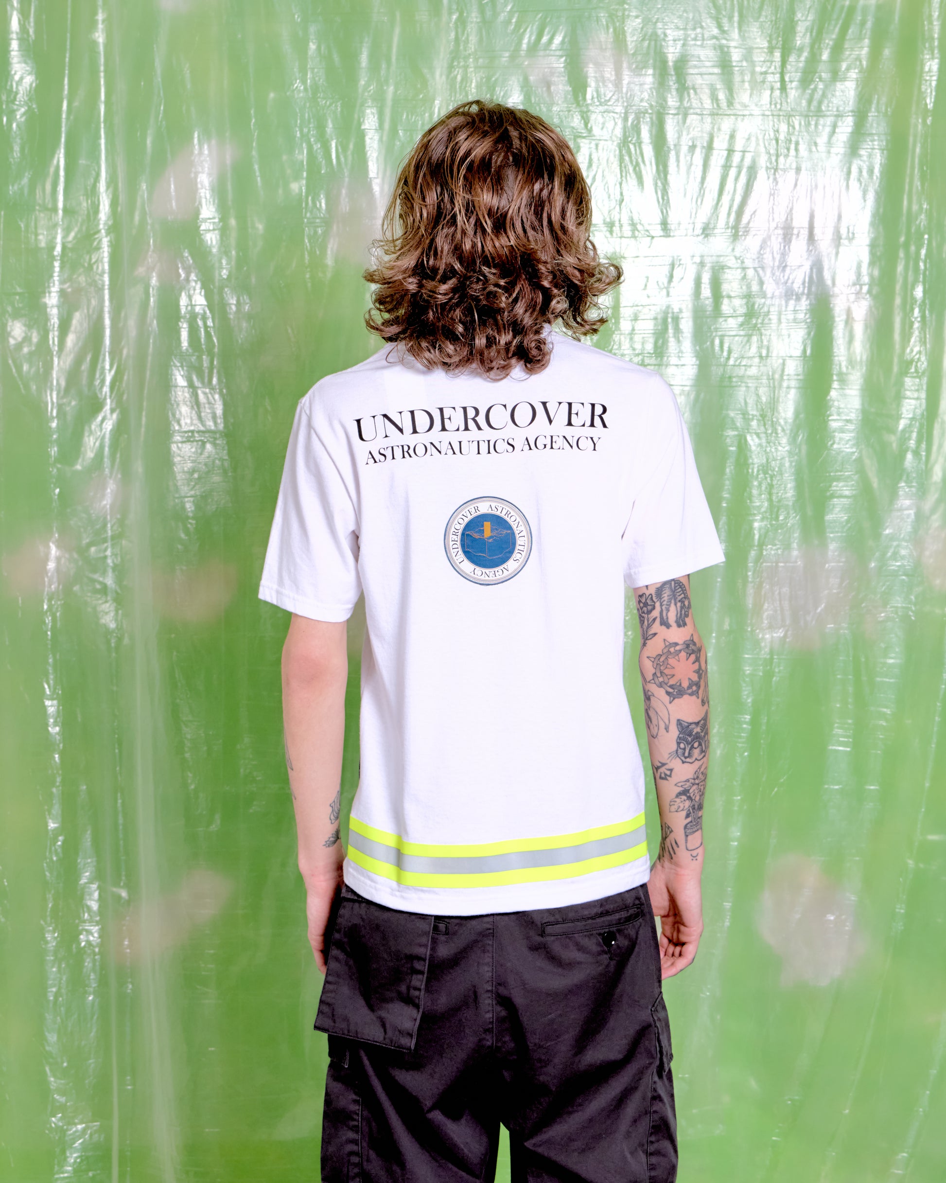 Person wearing a white 'Undercover Astronautics Agency' shirt with a logo on the back, standing against a green floral-patterned curtain.