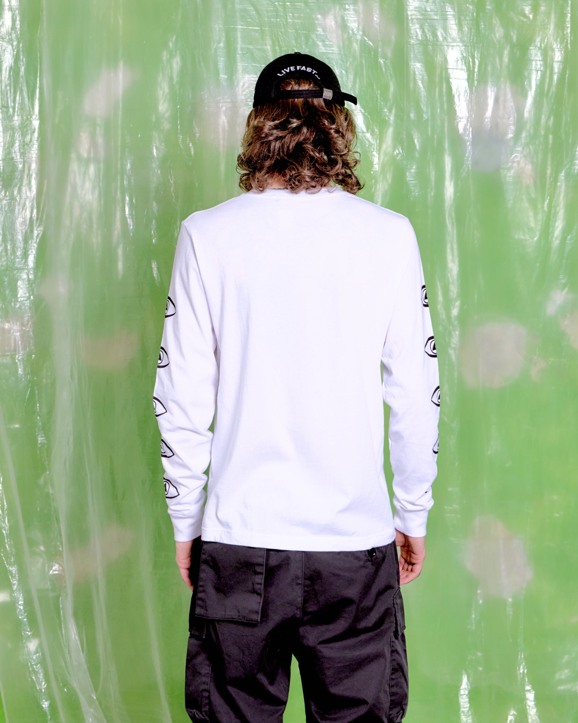 Person wearing a white long-sleeve shirt with black designs and dark pants against a green textured background