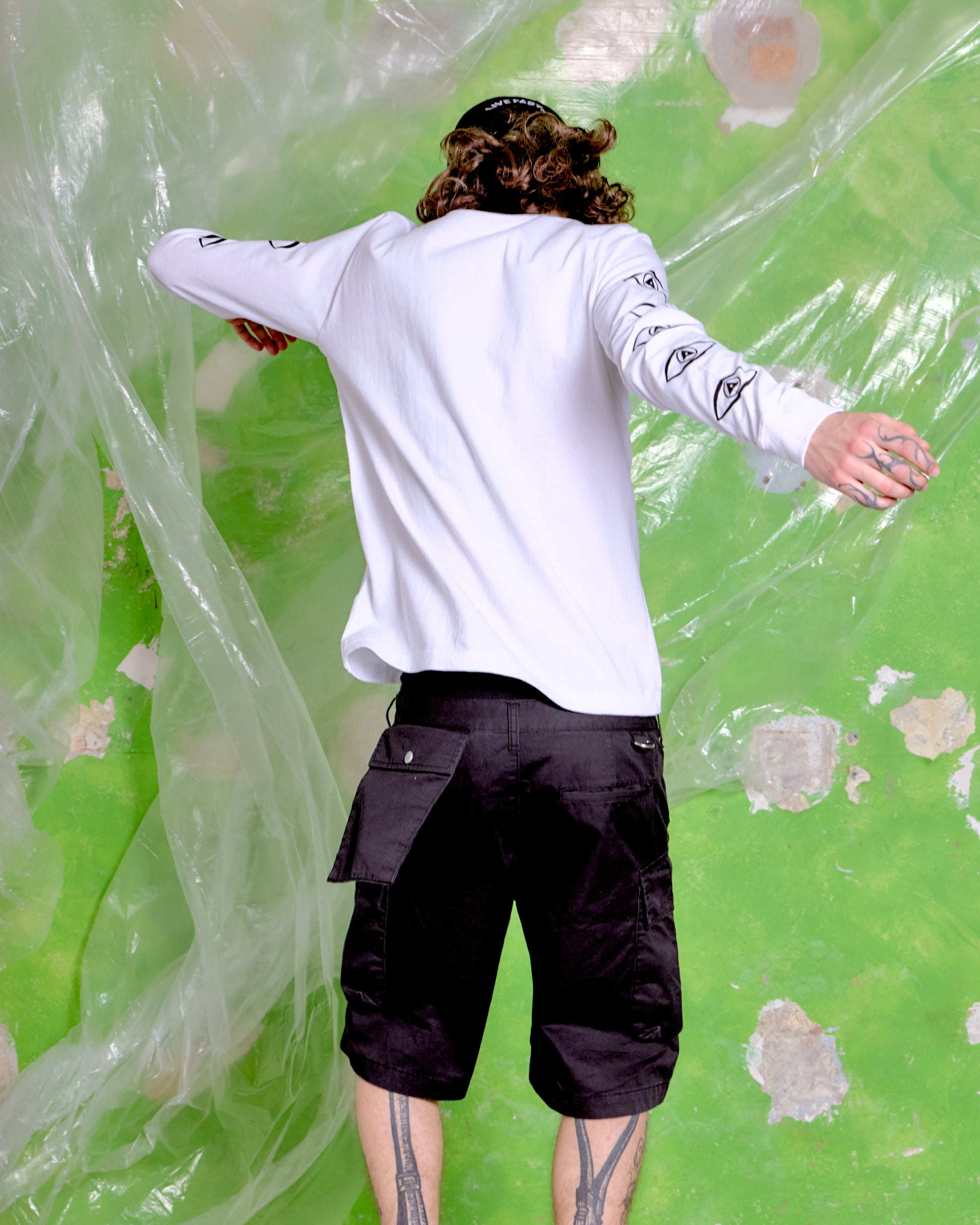 Person wearing a white long-sleeve shirt and black shorts standing in front of a green wall with floral patterns.