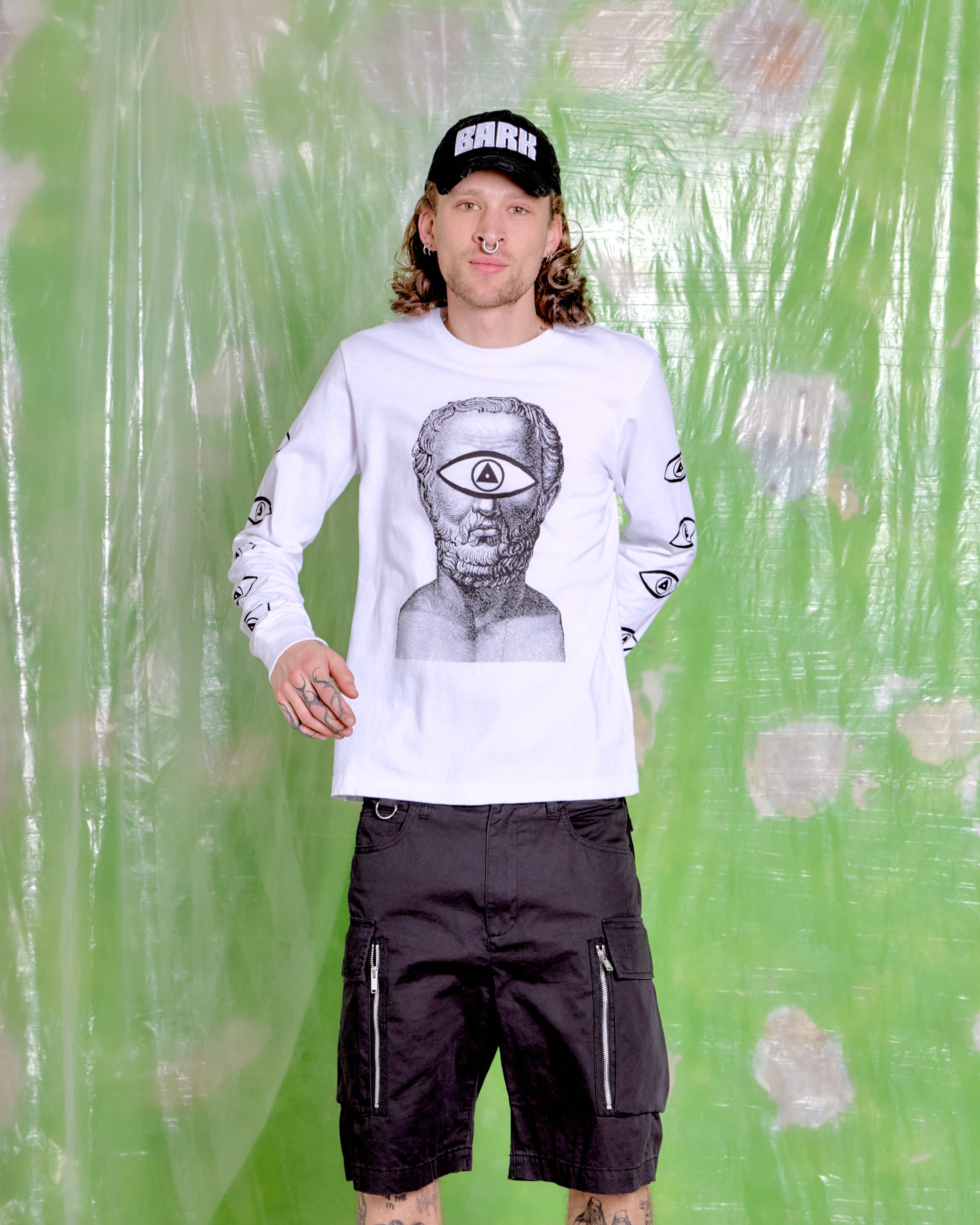 Person wearing a white long-sleeve shirt with graphic design and black cap, standing against a green textured background.
