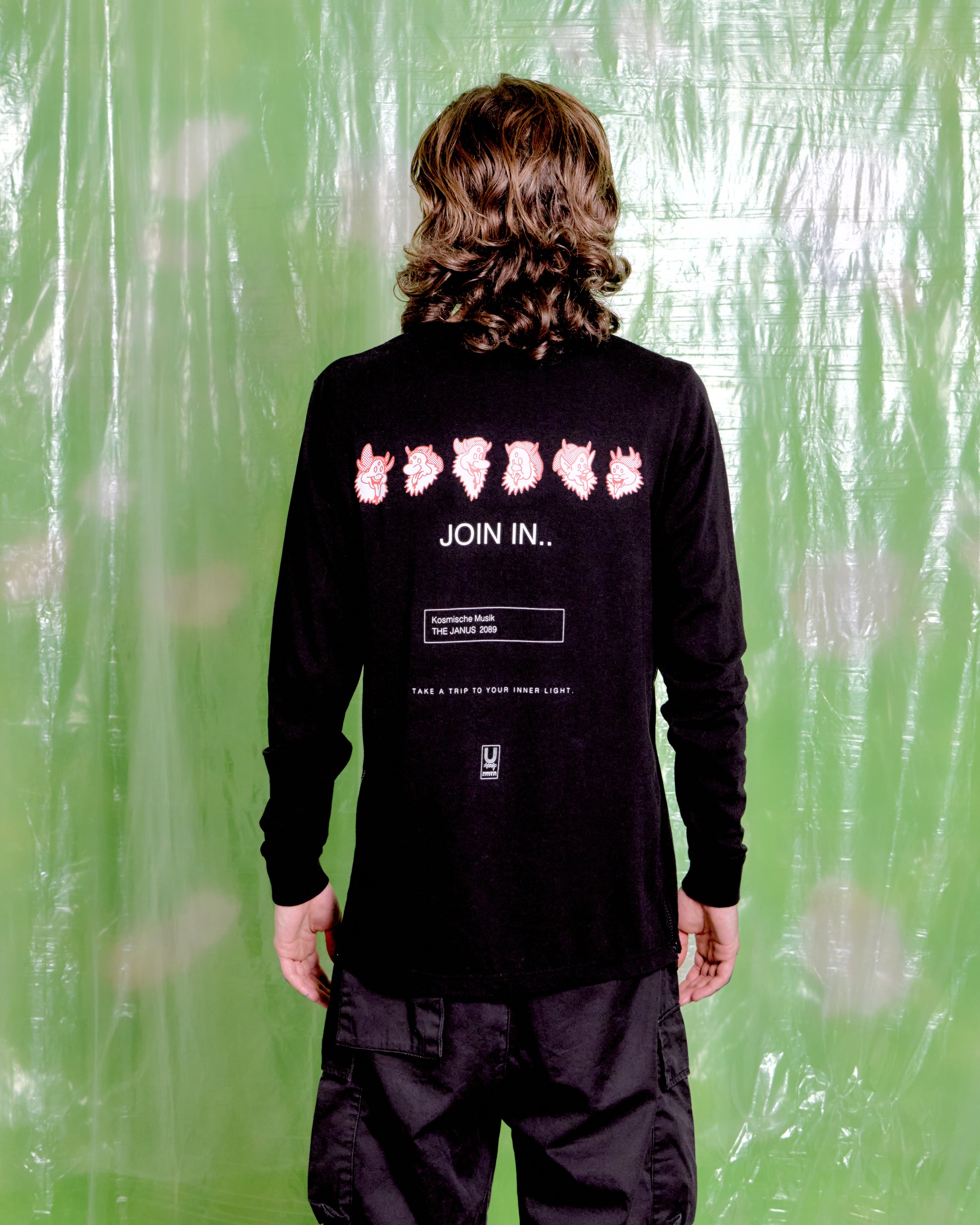 Person wearing a black long-sleeve shirt with pink graphics and text on the back, standing against a green curtain.