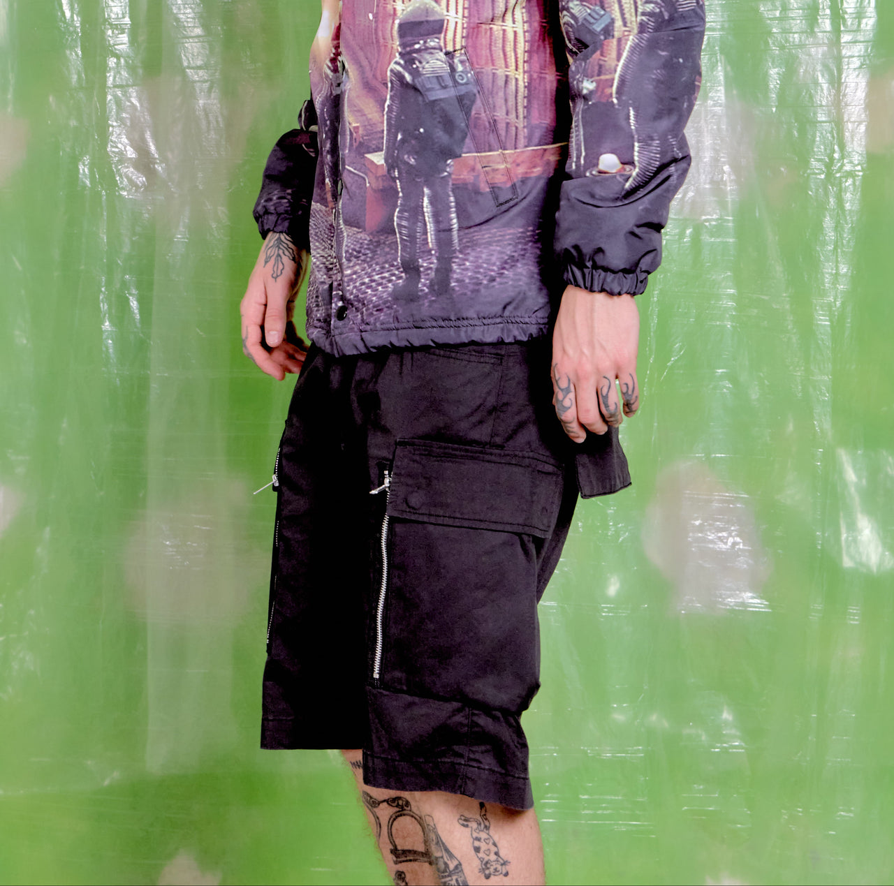 Person wearing black cargo shorts with zippers against a green curtain background