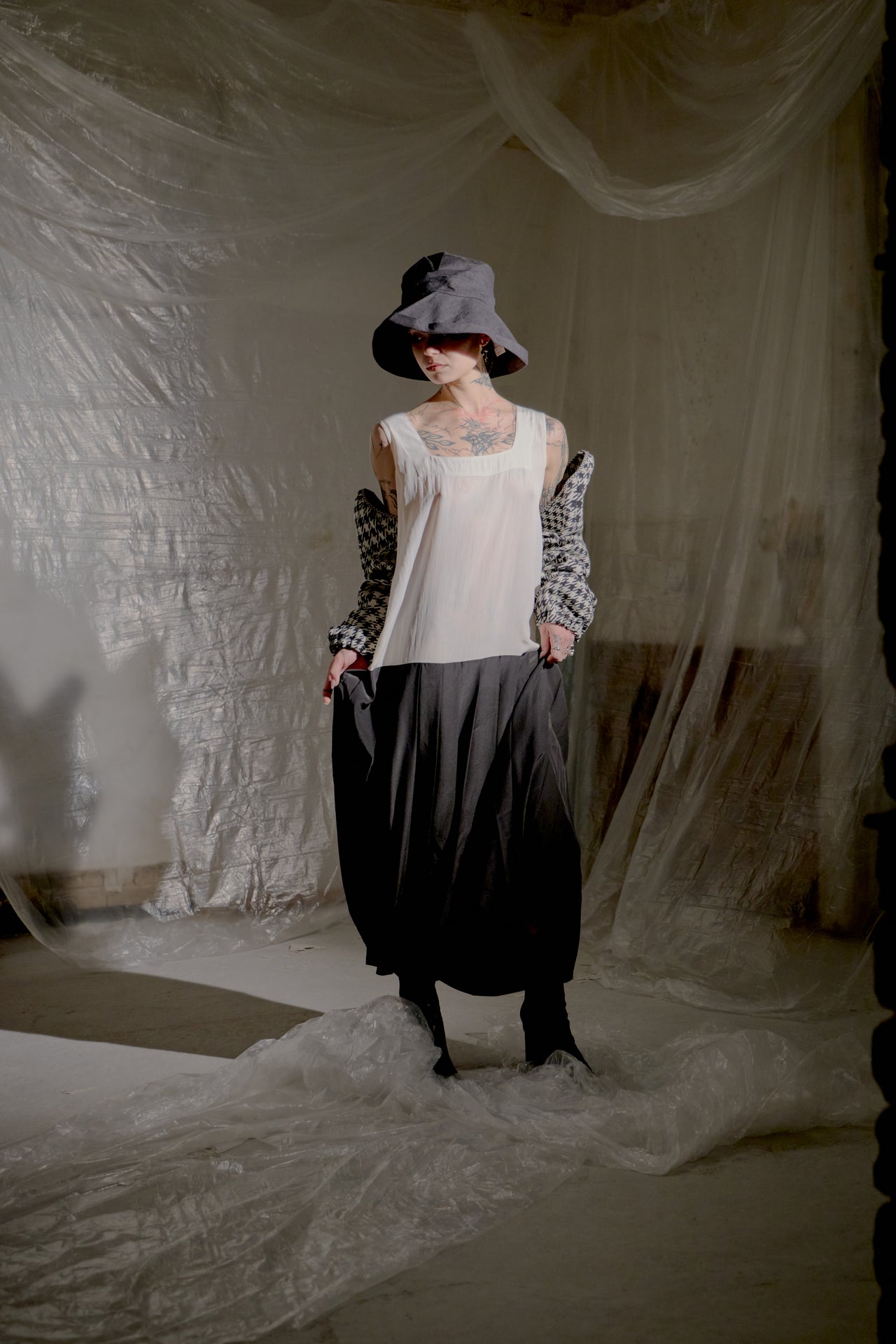Person wearing a black and white Limi Feu dress with a hat, standing in a softly lit room with draped fabric.