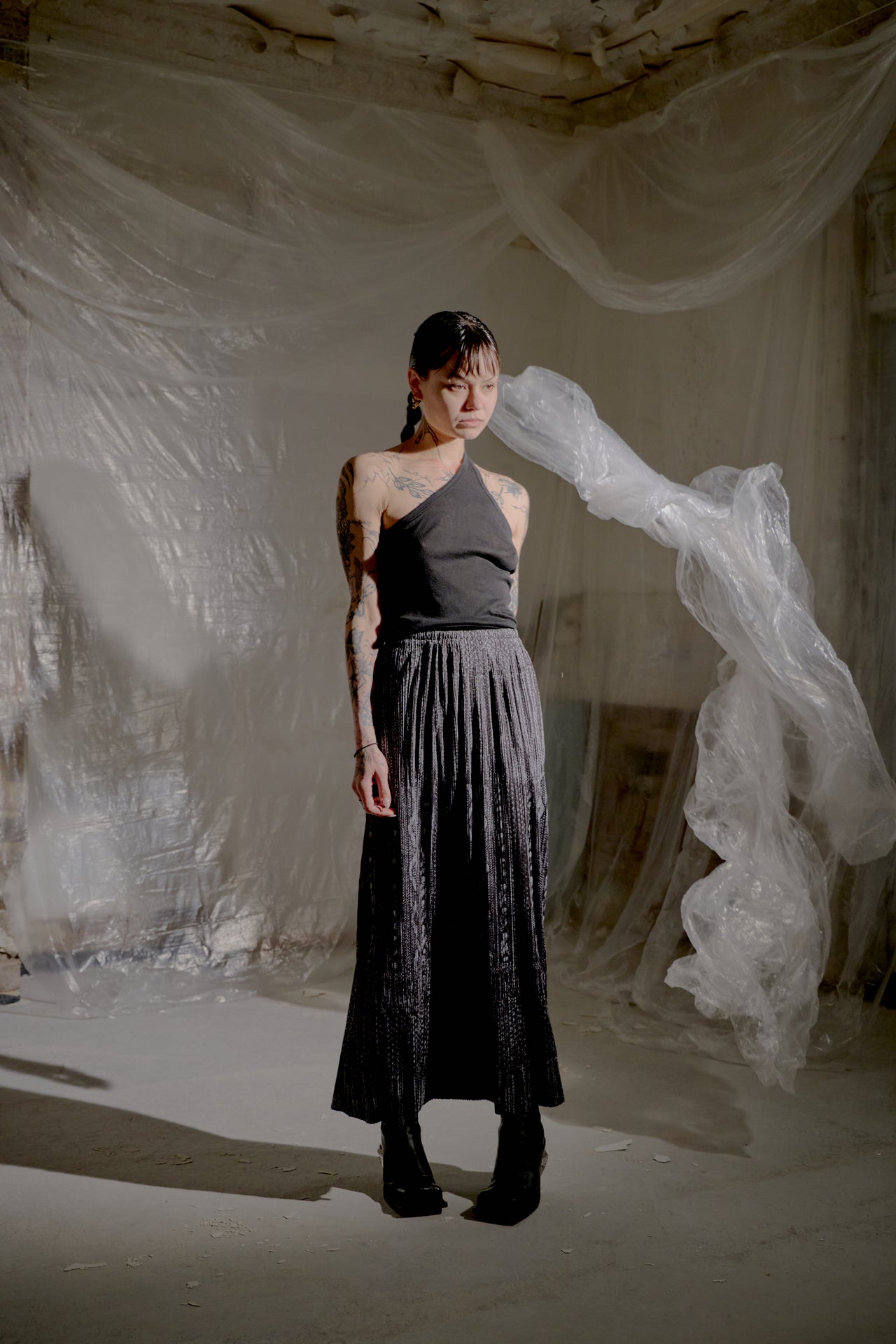 Person wearing a dark top and skirt standing in a textured, neutral-coloured room.