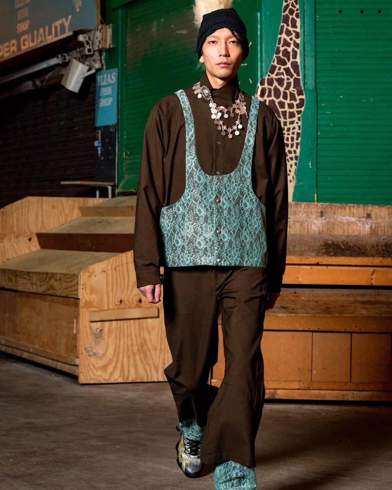 Person wearing a brown Kiko Kostadinov outfit with a teal patterned top, standing in front of a green wall with a giraffe graphic.