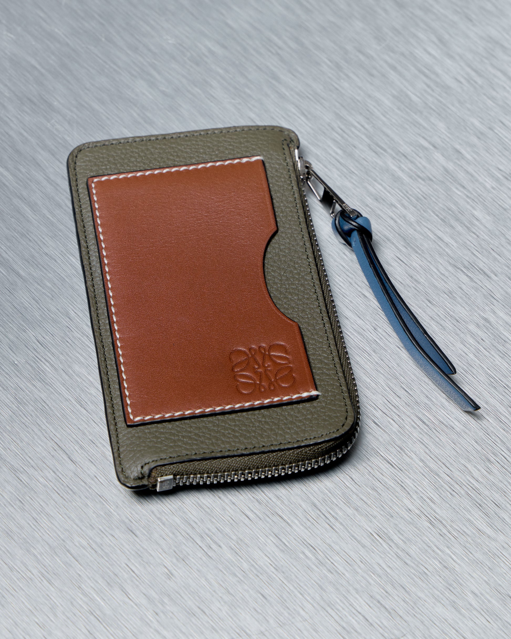 A textured Loewe leather wallet lies on a metal surface. The wallet has olive and brown sections with a zippered edge and a blue strap