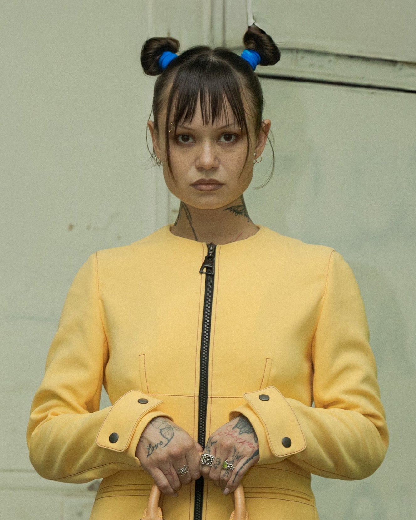 Person in a yellow Louis Vuitton coat holding a patterned bag in an urban setting with posters and an air conditioner.