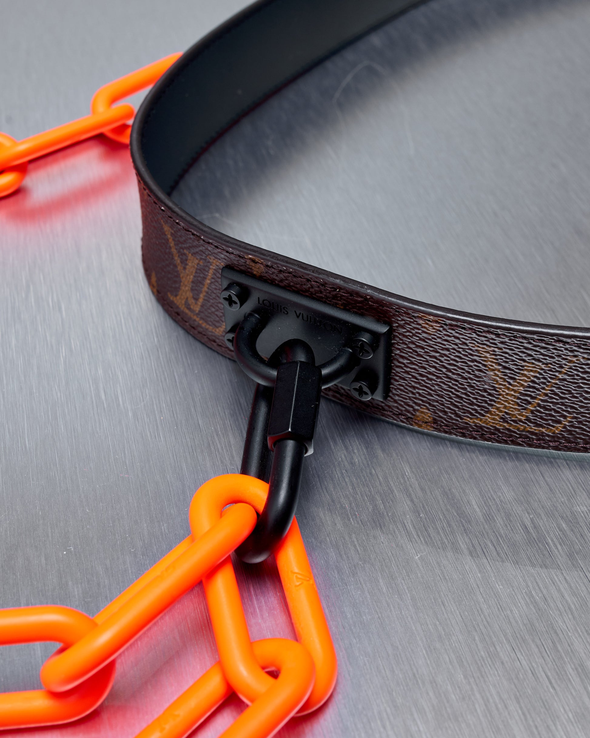 A brown leather Louis Vuitton strap connects to a vibrant orange plastic chain using a black metal clasp on a metallic surface