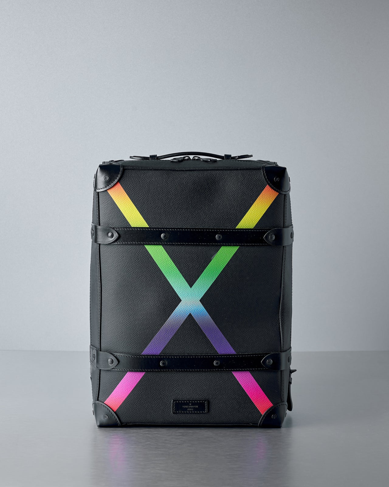 A sleek black Louis Vuitton suitcase with colourful rainbow gradient "X" on the front. It has black leather accents and a modern, stylish look, resting on a grey surface.