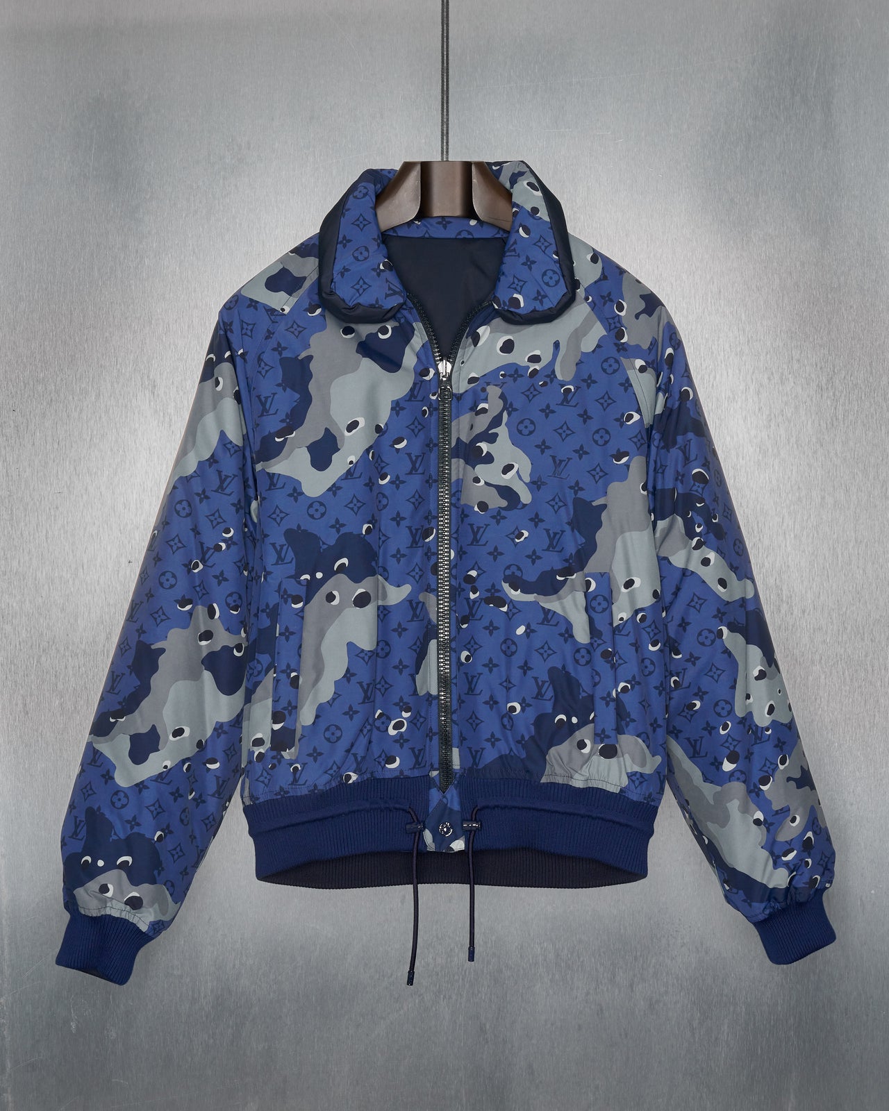 A blue camouflage padded Louis Vuitton jacket with a drawstring waist, ribbed cuffs, and snap button pockets, featuring silver branded hardware.