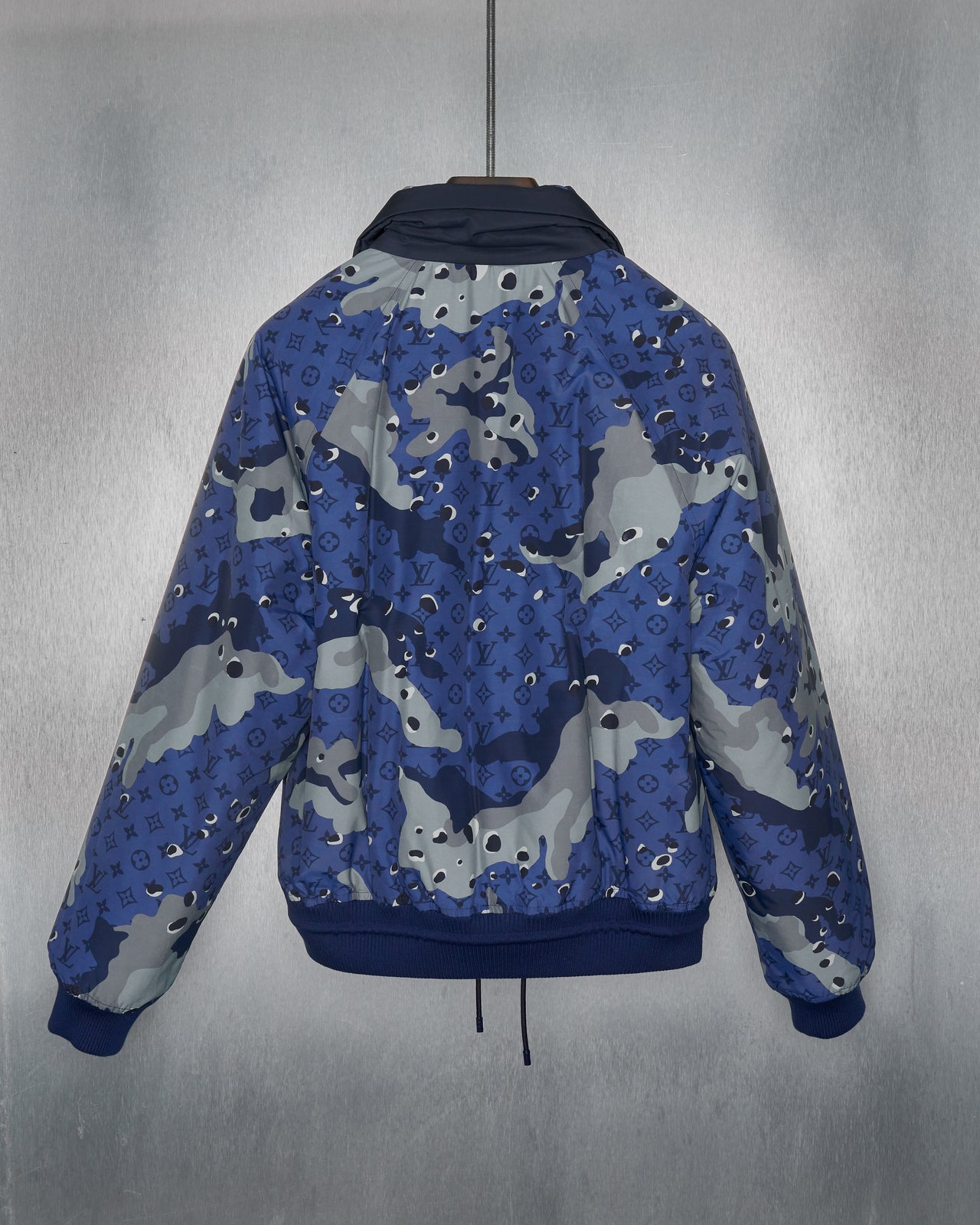 A blue camouflage padded Louis Vuitton jacket with a drawstring waist, ribbed cuffs, and snap button pockets, featuring silver branded hardware.