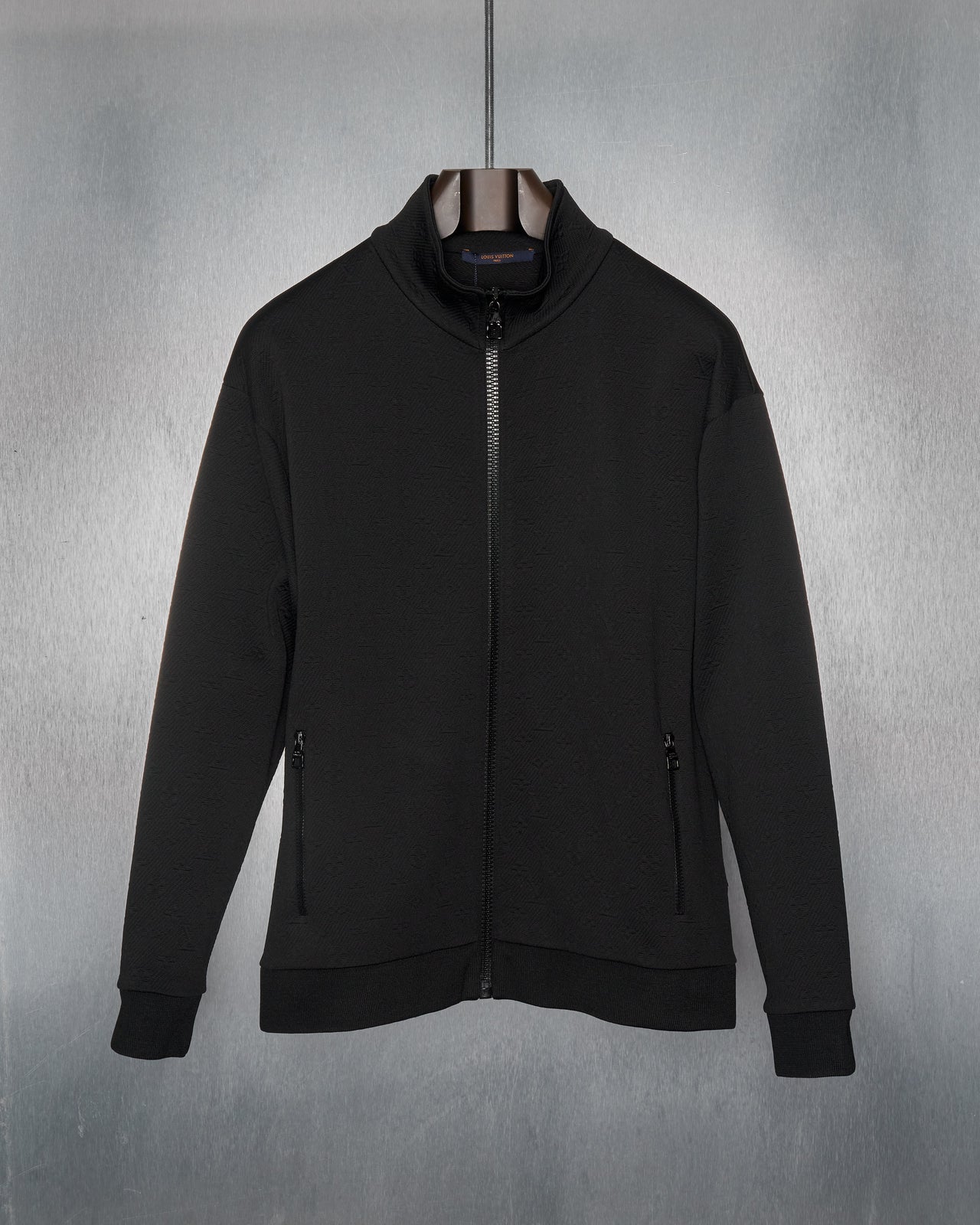 A black zip-up Louis Vuitton track jacket with monogram-embossed jacquard twill, featuring a high collar, zip pockets at the hips, ribbed cuffs and base. The jacket is displayed against a metallic background.