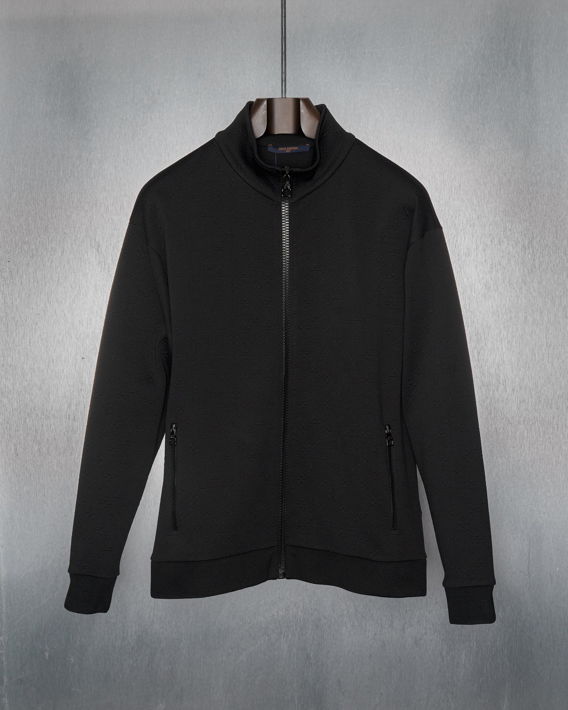 A black zip-up Louis Vuitton track jacket with monogram-embossed jacquard twill, featuring a high collar, zip pockets at the hips, ribbed cuffs and base. The jacket is displayed against a metallic background.