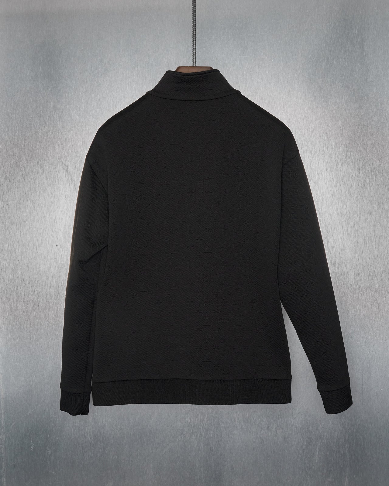 A black zip-up Louis Vuitton track jacket with monogram-embossed jacquard twill, featuring a high collar, zip pockets at the hips, ribbed cuffs and base. The jacket is displayed against a metallic background.
