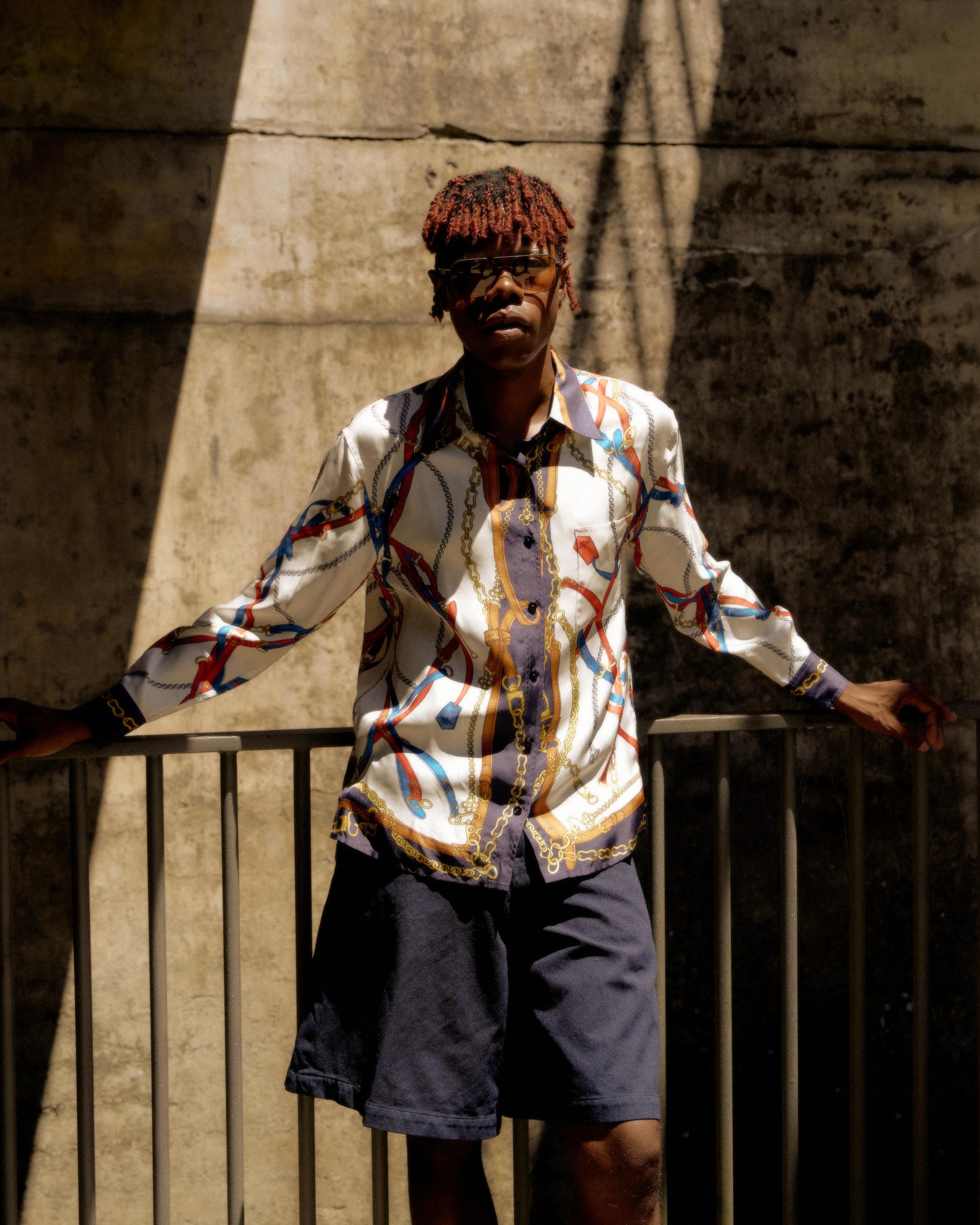 A person stands confidently against a textured concrete wall, wearing sunglasses, a colorful patterned Louis Vuitton shirt, and navy shorts. Sunlight casts dramatic shadows.