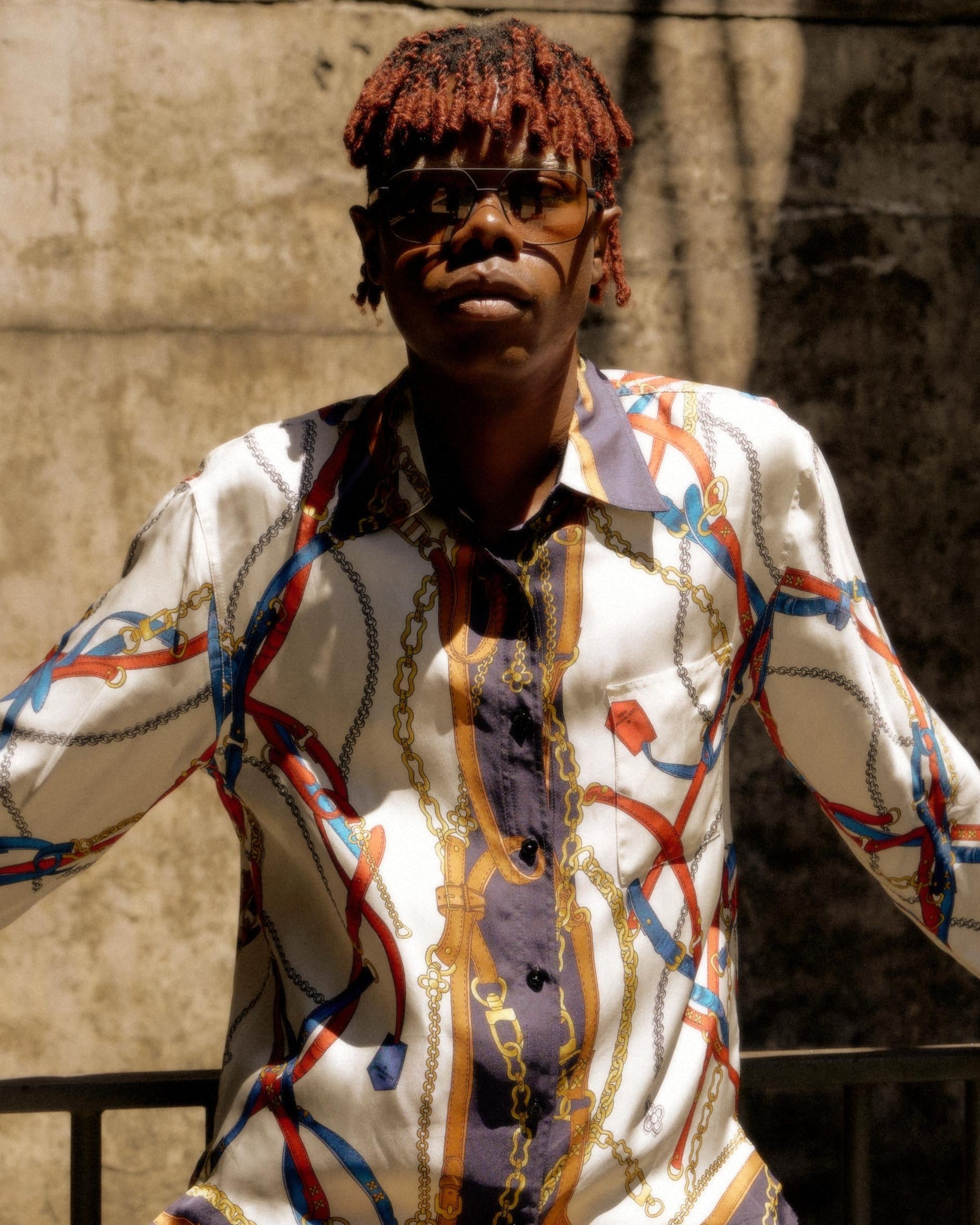 A person with short, red-tinted hair and sunglasses stands confidently, wearing a patterned Louis Vuitton shirt with chains and ropes. A textured wall is in the background.