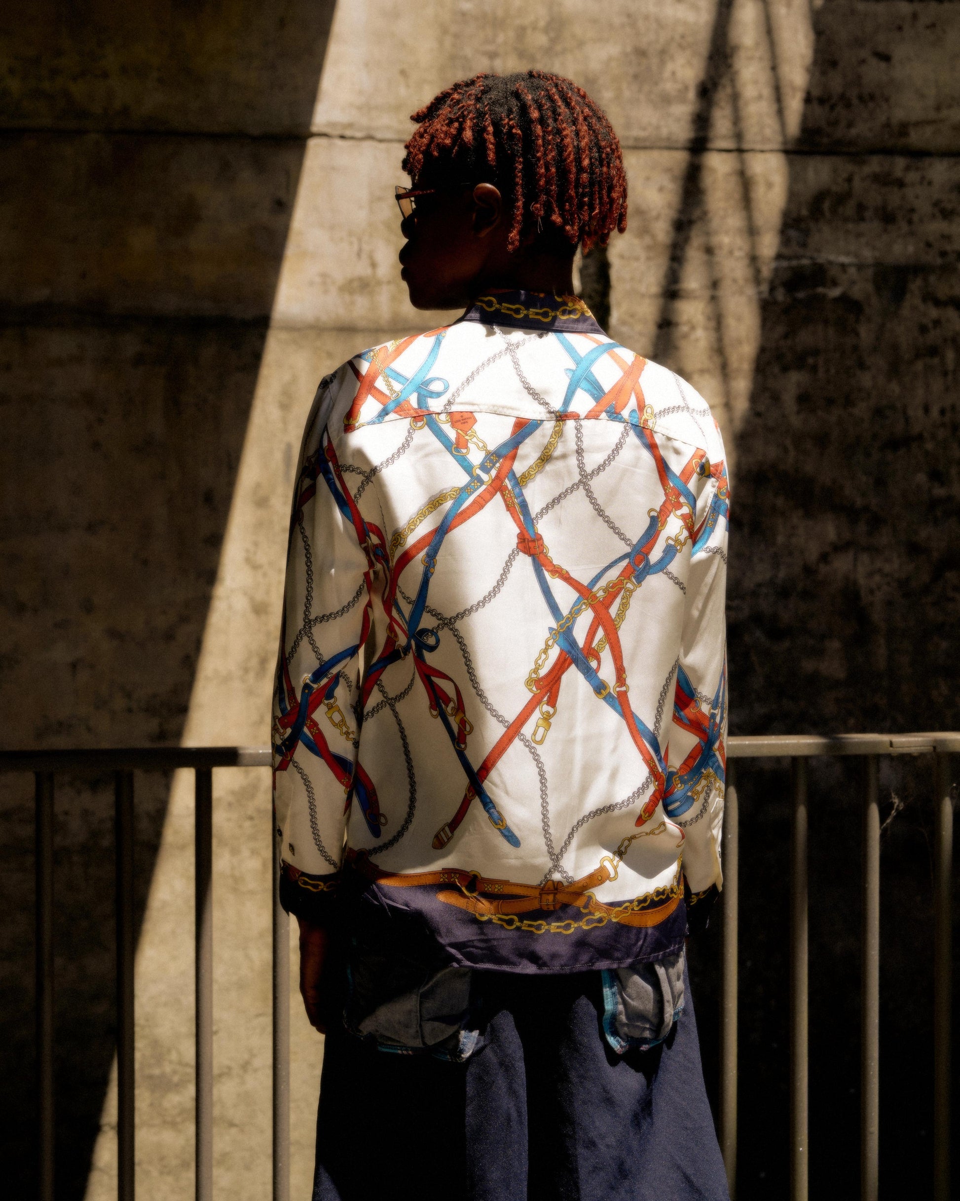 A person stands in shadow against a concrete wall, wearing a colorful, chain-patterned Louis Vuitton shirt and blue pants. The mood is contemplative and stylish.
