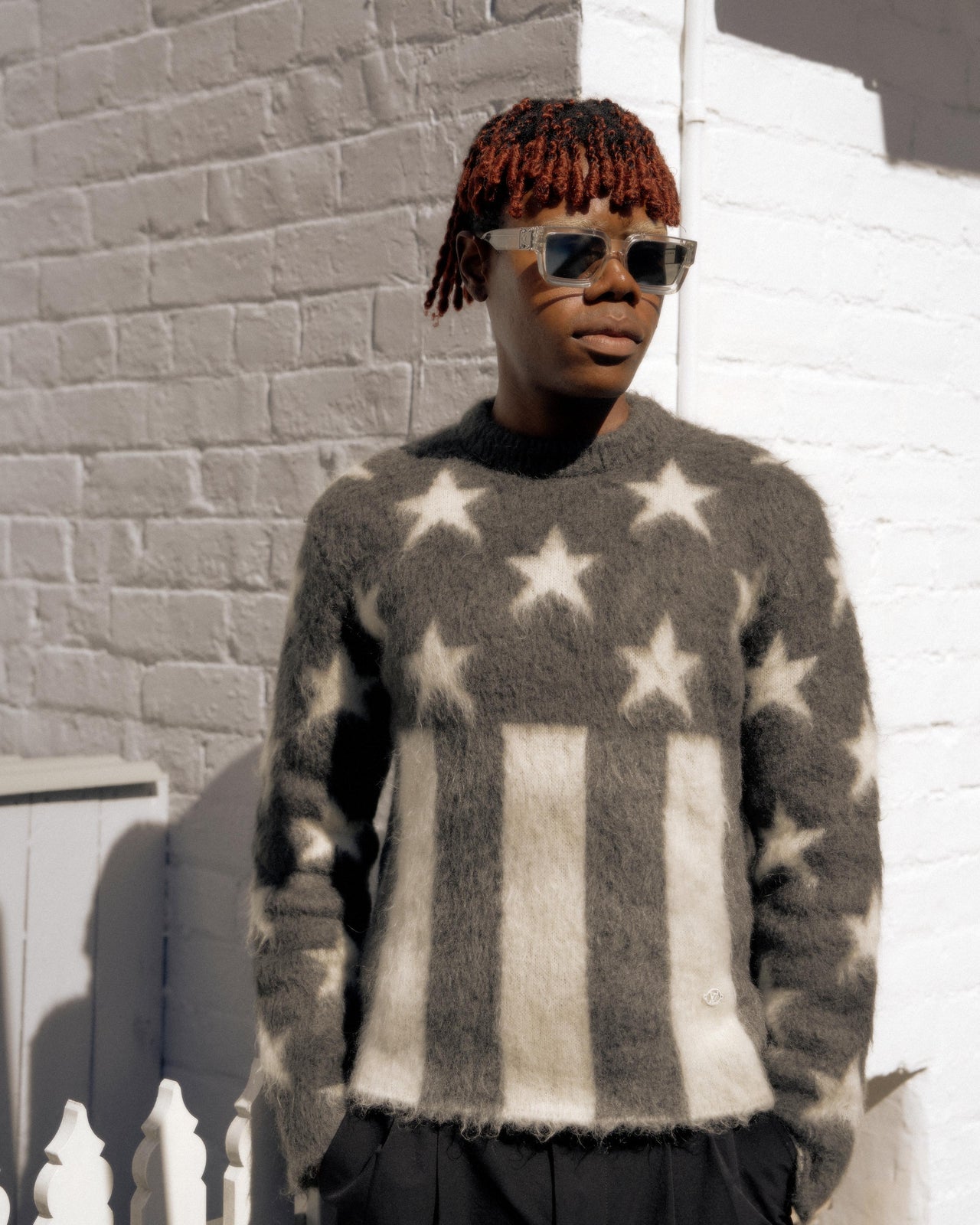 Model walking Louis Vuitton runway in a relaxed, confident manner, wearing a fuzzy sweater with stars and stripes and a flowing grey pleated skirt. The background is softly lit.