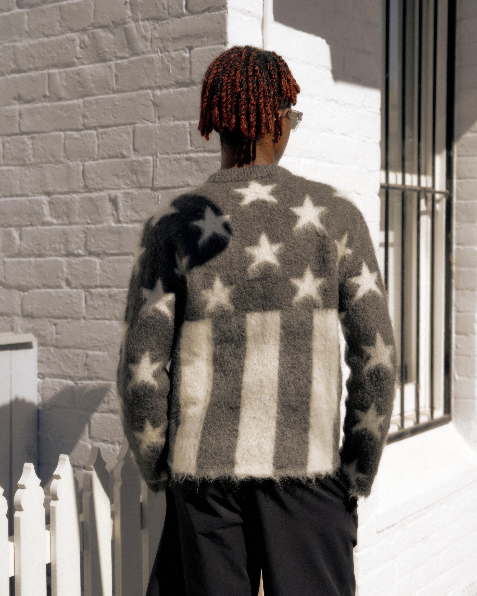 A person with red braided hair wearing a fuzzy Louis Vuitton sweater with an American flag pattern stands outside against a sunlit white brick wall, conveying a casual vibe.