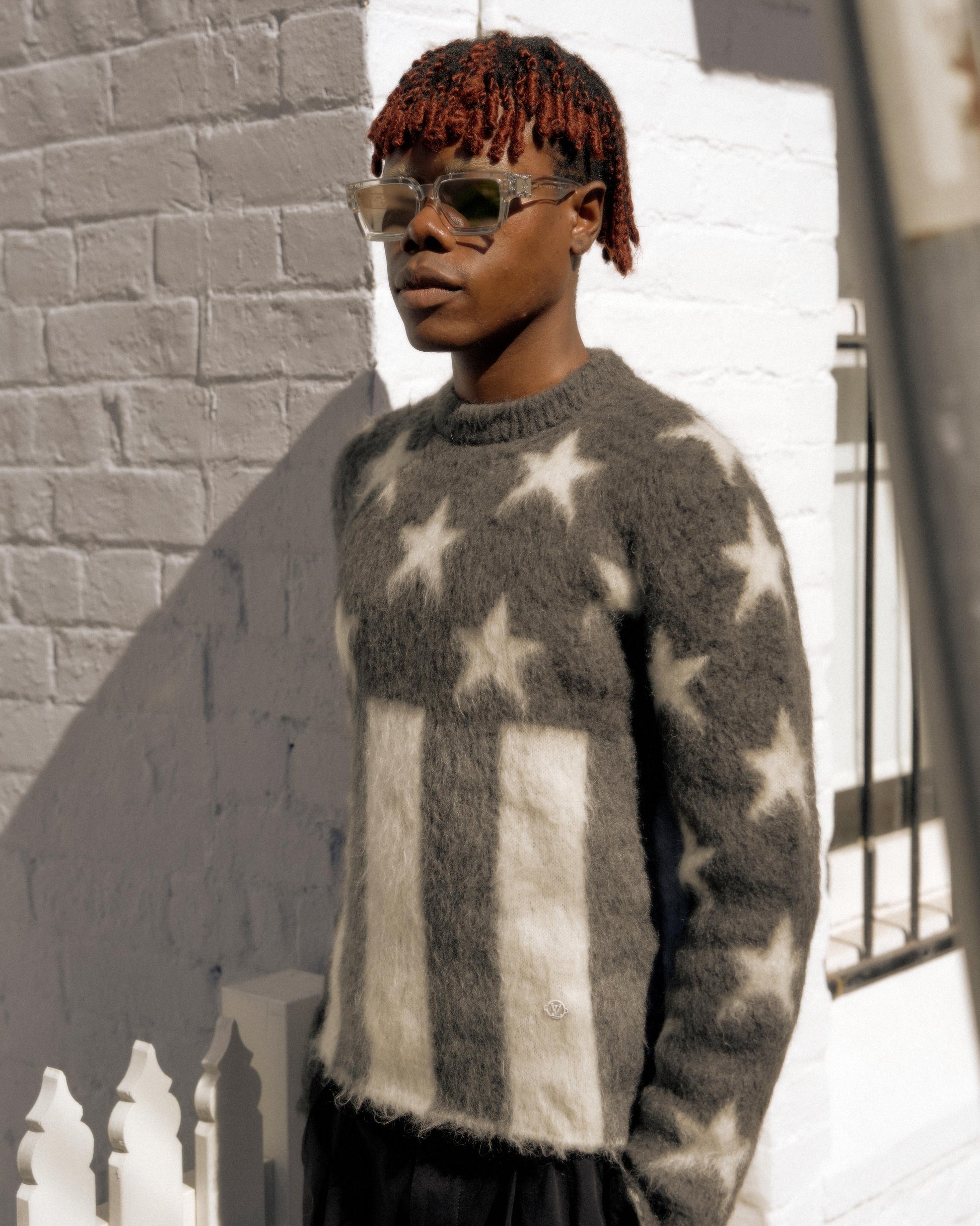 A person stands against a sunlit brick wall wearing a fuzzy Louis Vuitton sweater with white stars and rectangular patterns, and sunglasses, projecting a calm, stylish vibe.