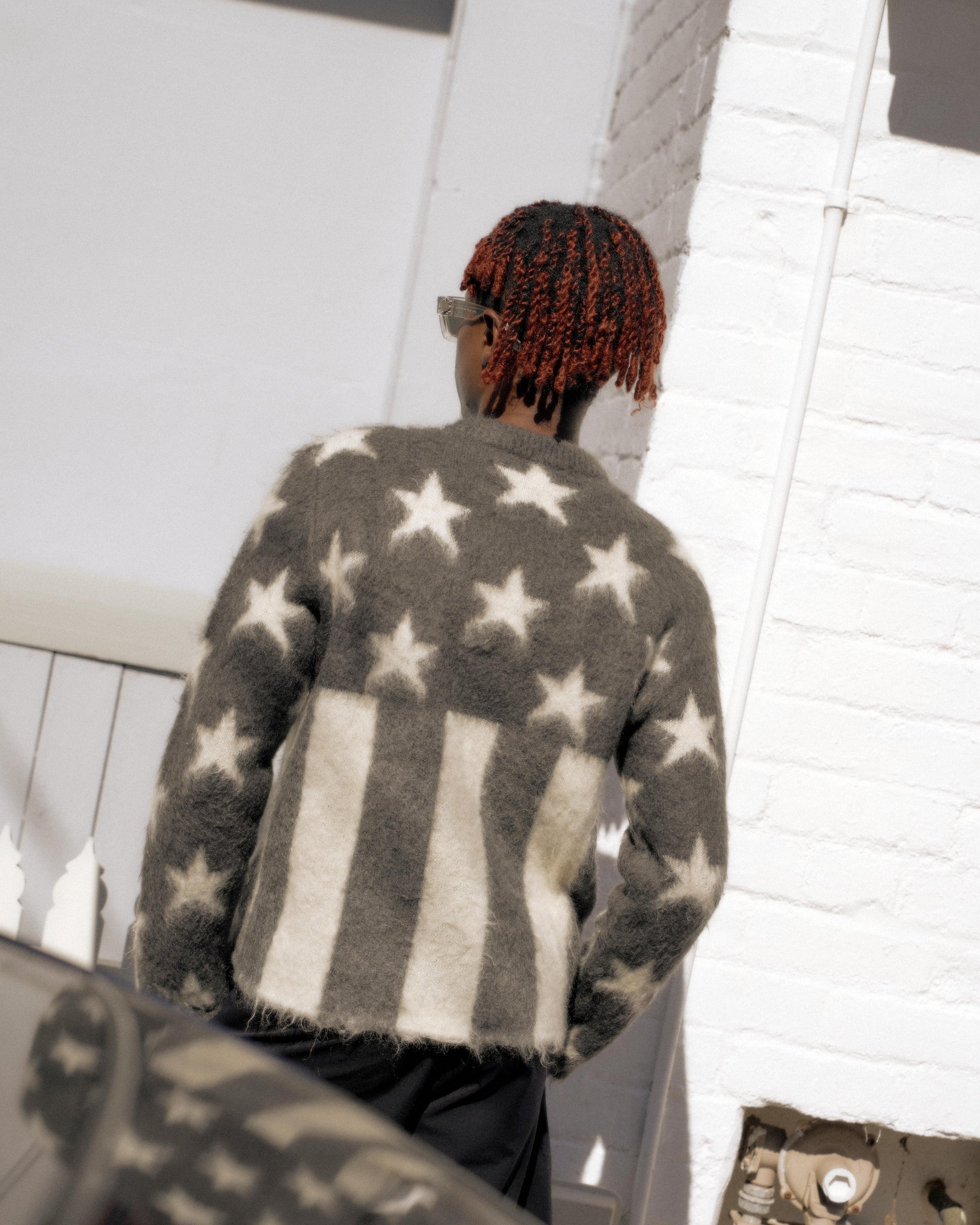 A person with red braided hair wears a fuzzy Louis Vuitton sweater with star and stripe patterns, reminiscent of the U.S. flag, standing near a white brick wall.