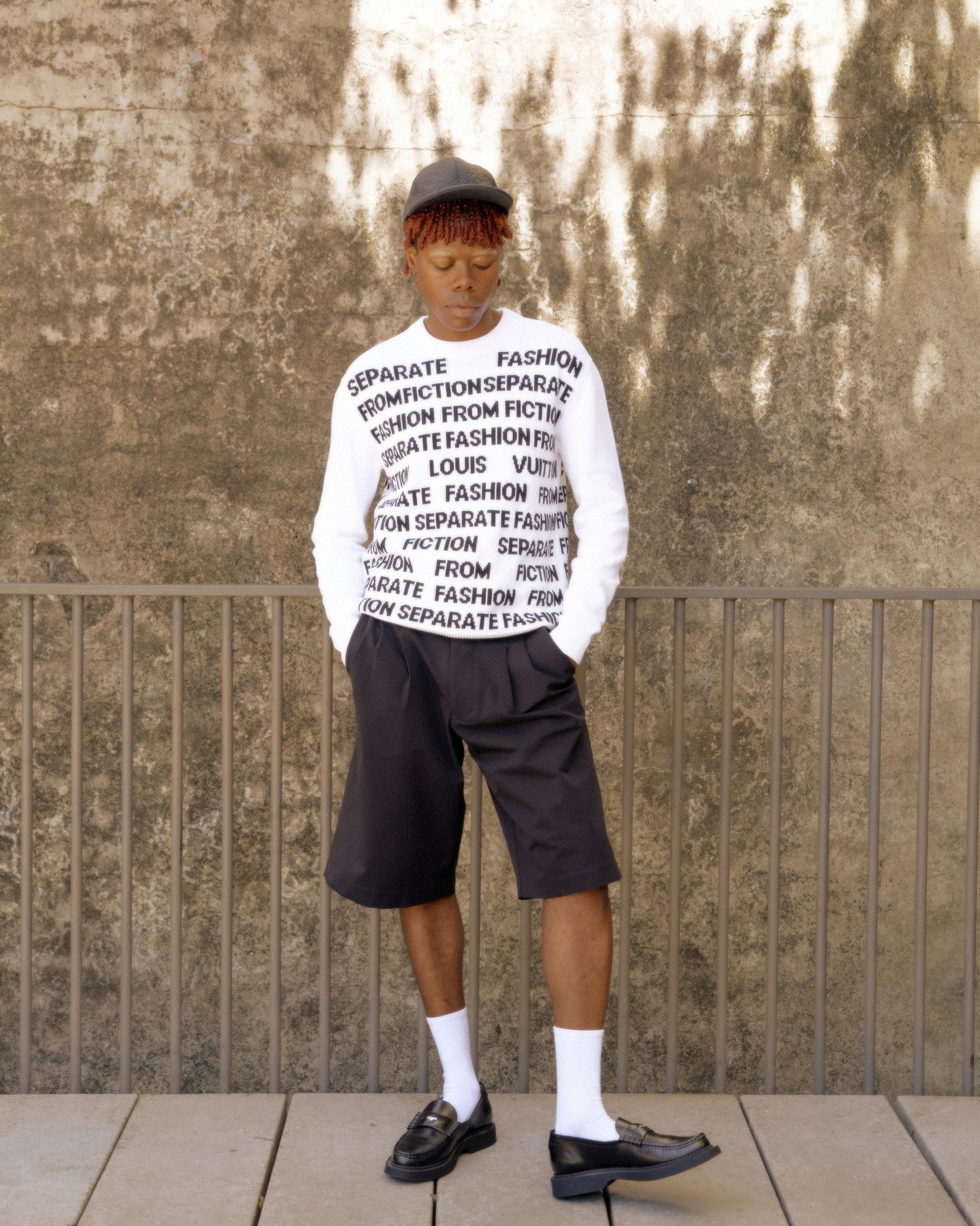 A person stands against a textured wall, wearing a white Louis Vuitton sweater with black text, black shorts, white socks, and black loafers. The mood is contemplative. 