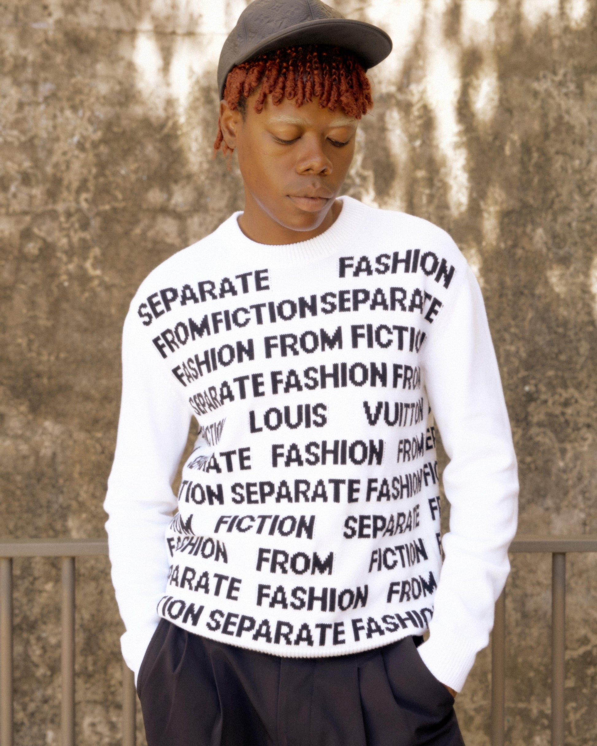 A person with red braids and a black cap wears a white Louis Vuitton sweater with black text that reads "SEPARATE FASHION FROM FICTION." The background is a textured wall.