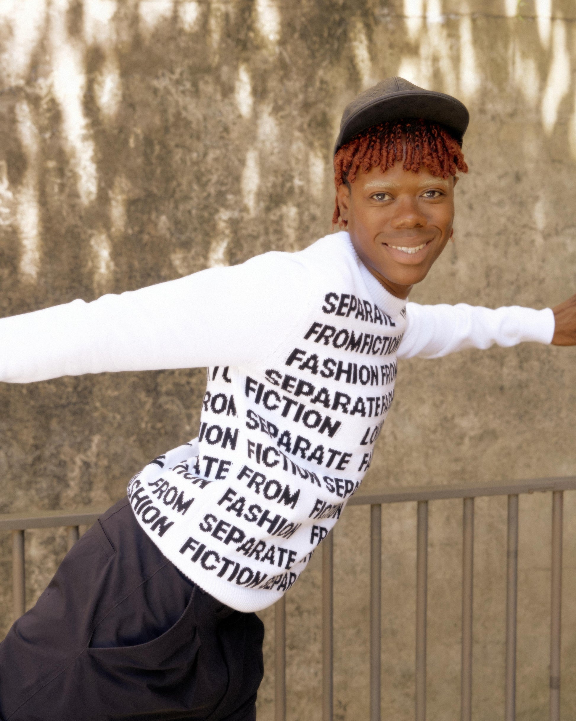 Person with red hair and a black cap poses joyfully, arms outstretched. Wears a Louis Vuitton sweater with "SEPARATE FASHION FROM FICTION" text. Background: textured wall.