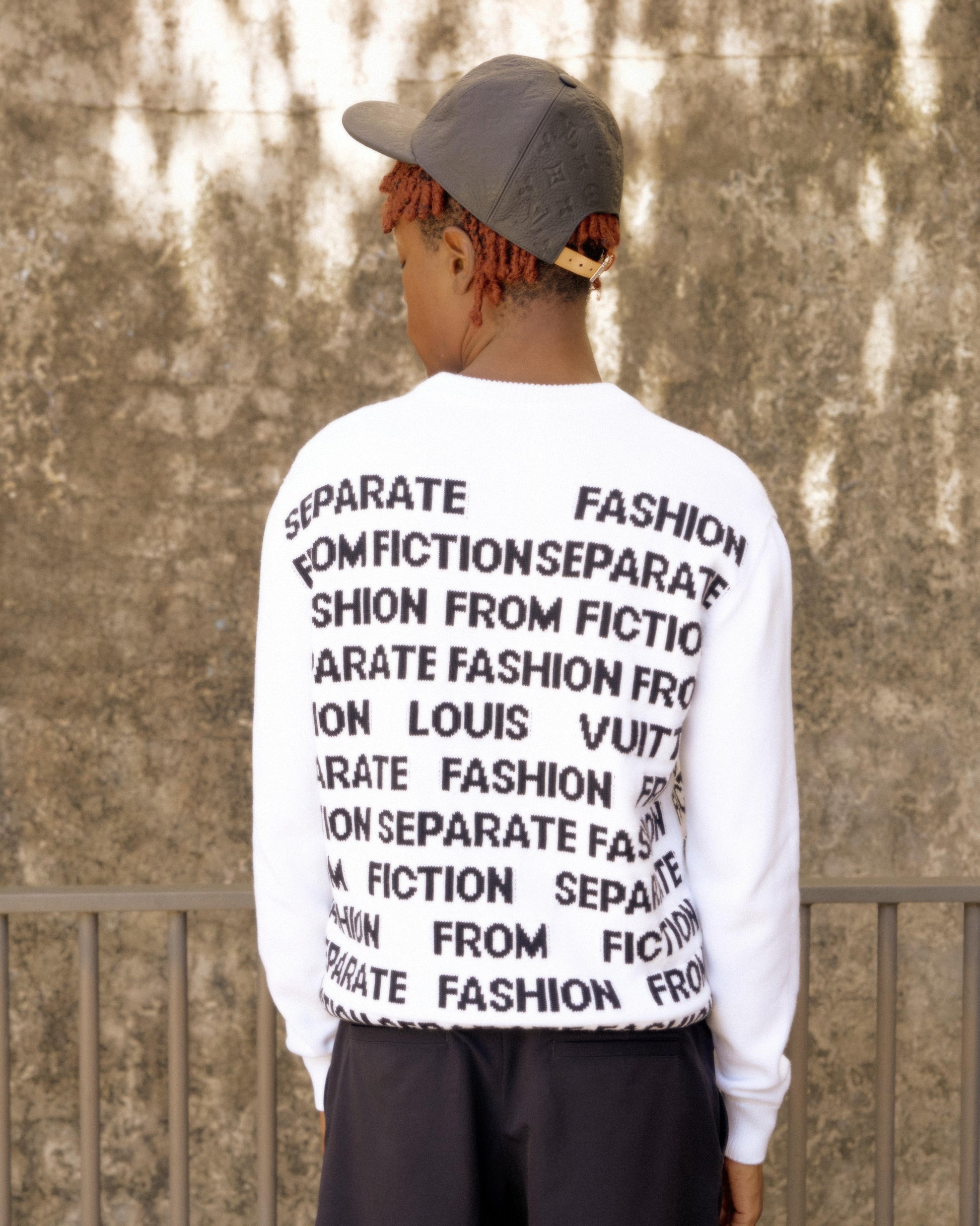 A person with red hair, wearing a gray cap and white Louis Vuitton sweater printed with "Separate Fashion from Fiction," stands against a textured wall, creating a thoughtful tone.