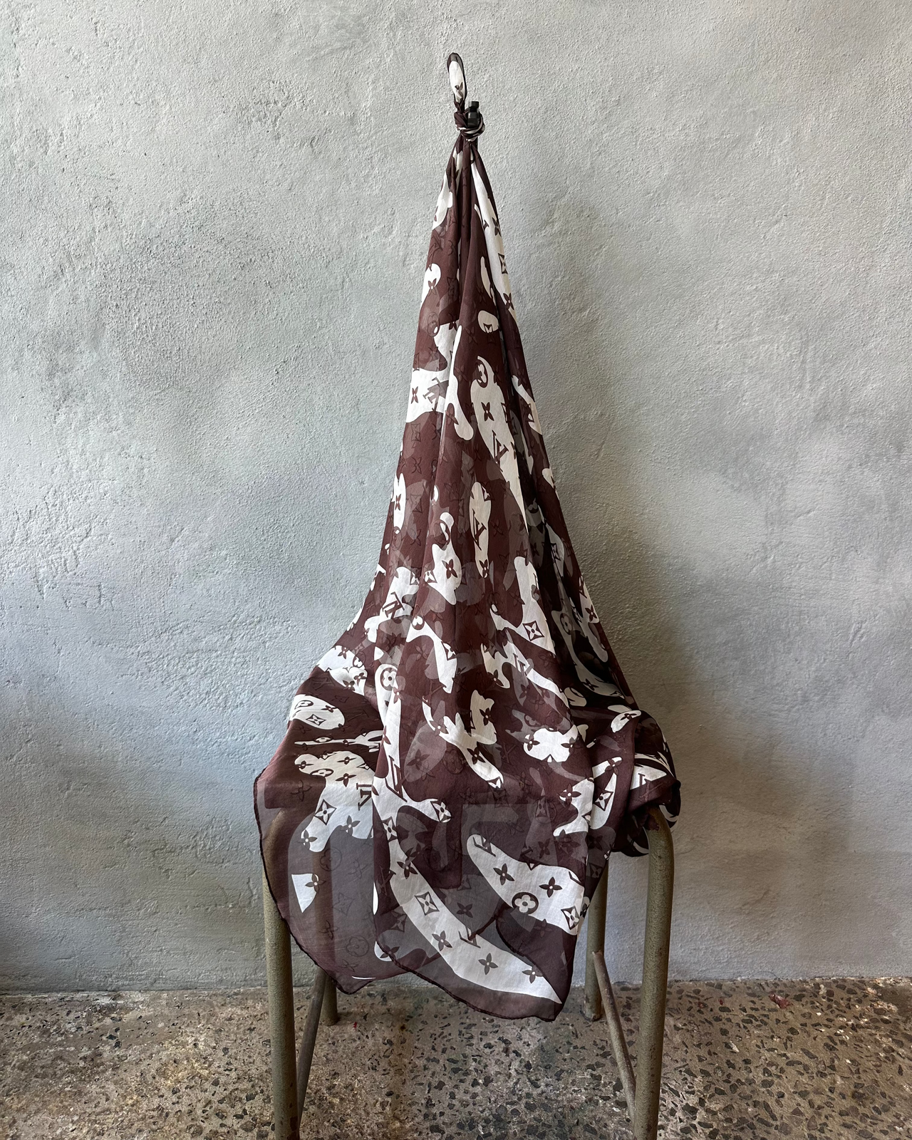 Brown patterned Louis Vuitton scarf hanging on a metal stand against a textured wall.
