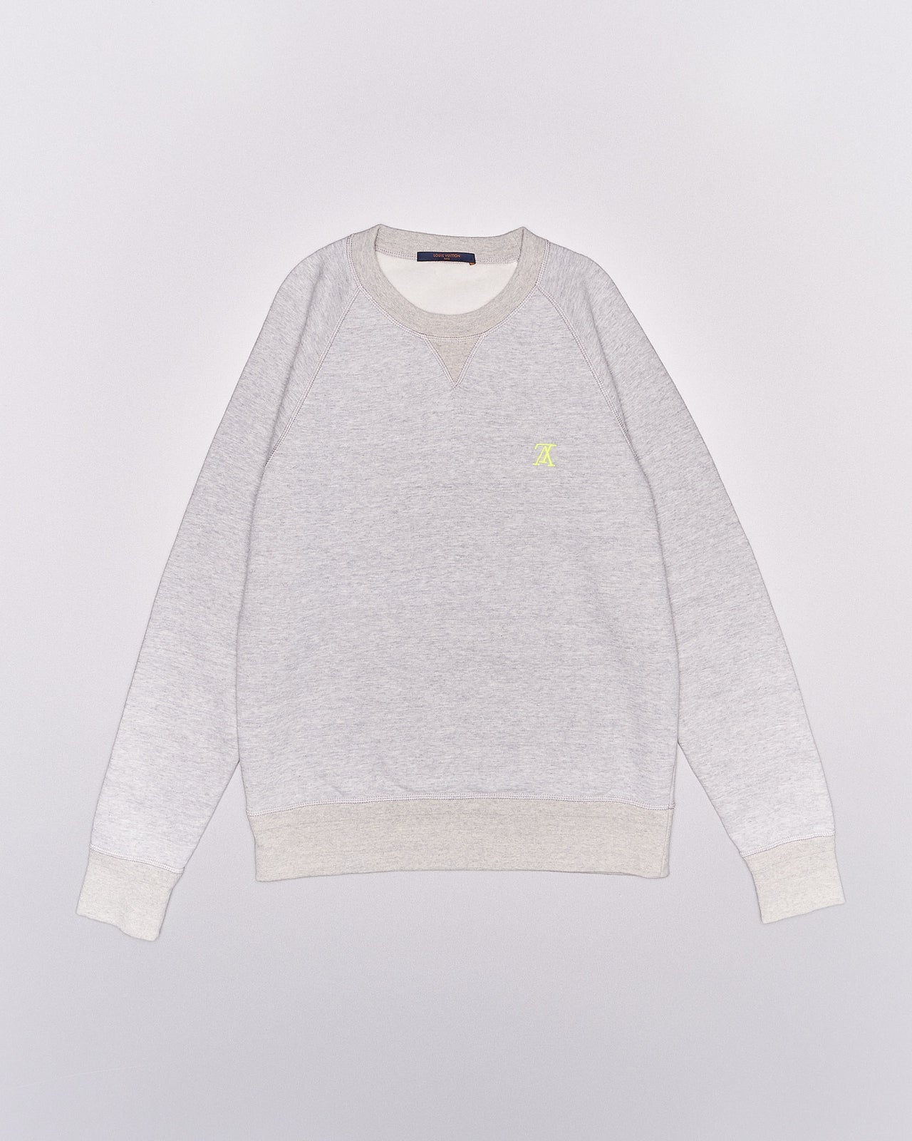 A light grey Louis Vuitton sweatshirt with beige ribbed cuffs and collar. It features a small yellow embroidered logo on the chest, conveying a casual and cozy feel.