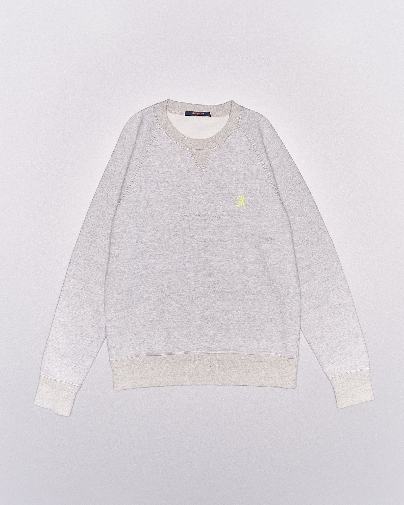 A light grey Louis Vuitton sweatshirt with beige ribbed cuffs and collar. It features a small yellow embroidered logo on the chest, conveying a casual and cozy feel.