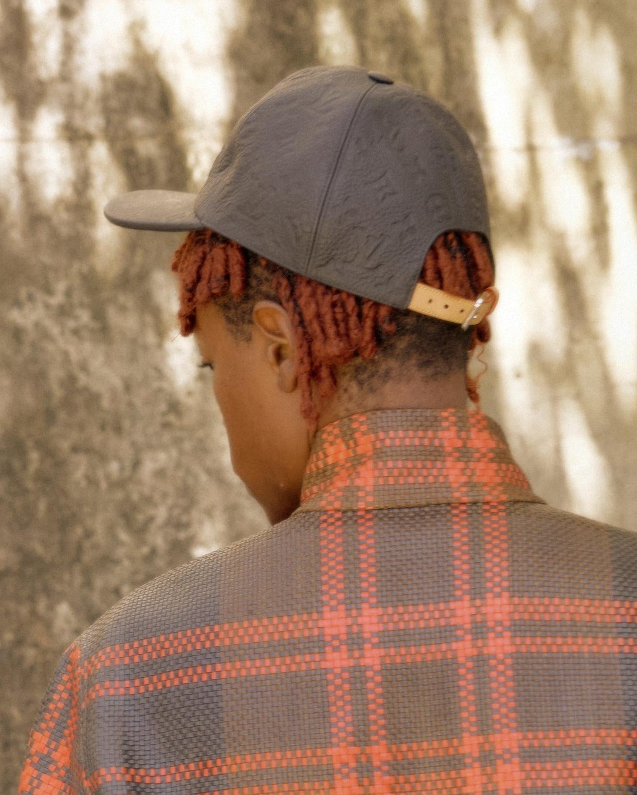 A person wearing a grey and orange Louis Vuitton checkered cap with a leather embossed monogram 1.1 cap on a textured background.