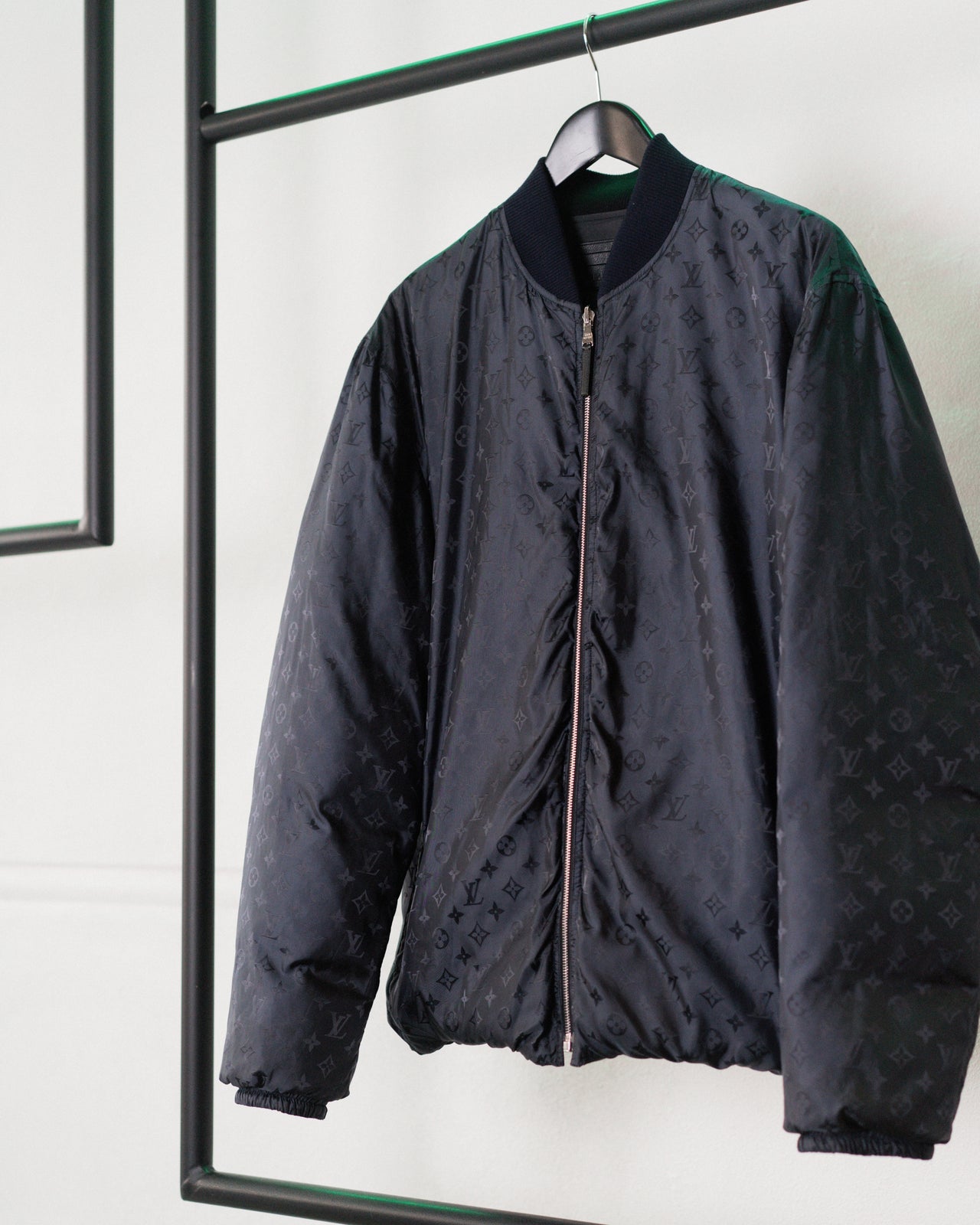 A Louis Vuitton down jacket with a monogram pattern, displayed on a hanger. The jacket is navy in color, has a ribbed collar, and features front zip closure with leather pull.