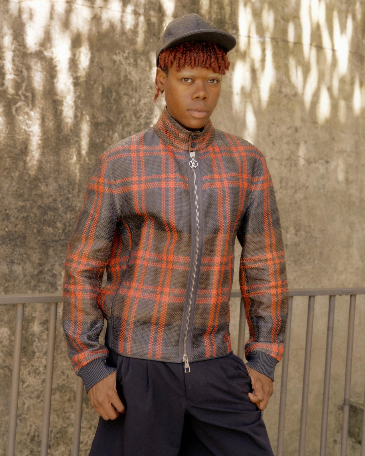 A male fashion model walks down an outdoor runway wearing a red and blue plaid Louis Vuitton jacket, checkered pants, a brown crossbody bag, and sandals, with an audience seated along the sides.