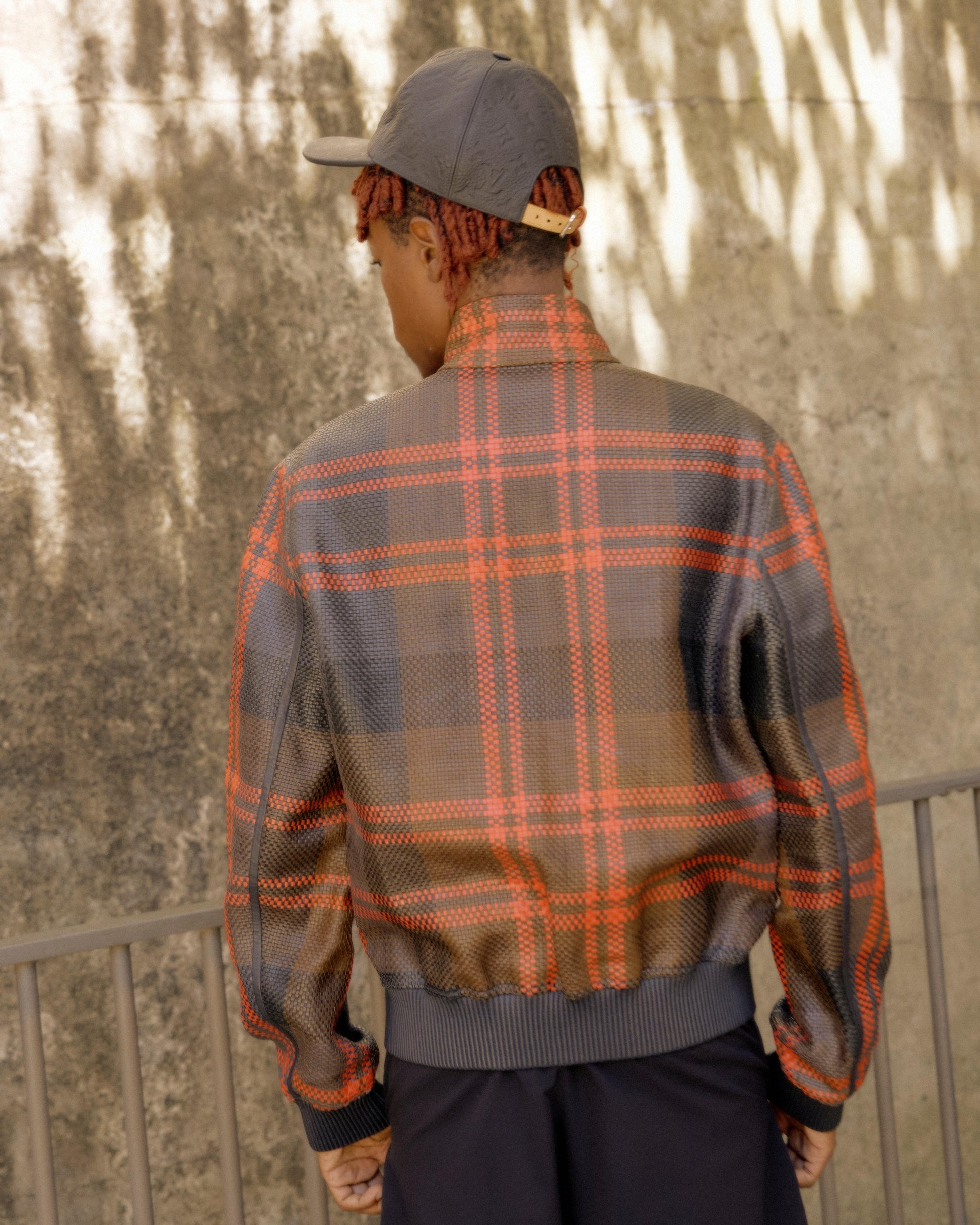A person with braided hair wearing a grey cap and a plaid Louis Vuitton jacket with red and brown tones stands facing a textured, sunlit wall outdoors.
