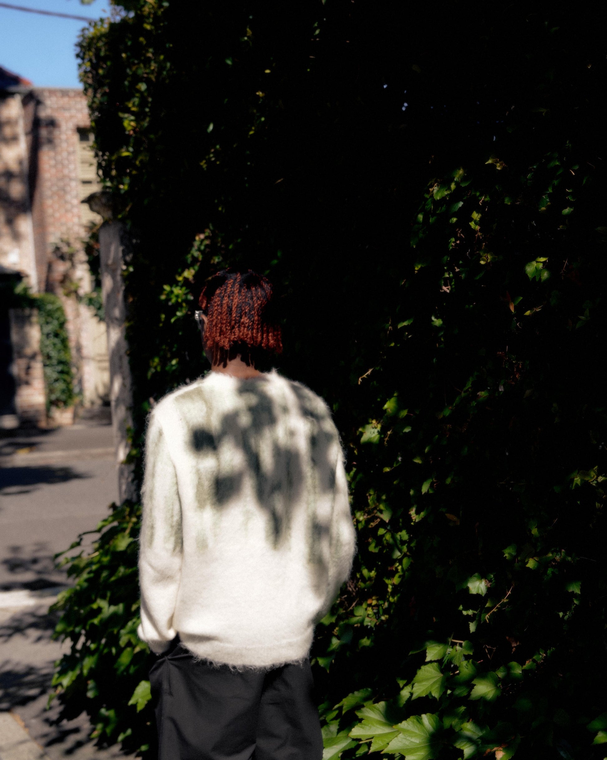 A person with red hair stands facing away, wearing a white fuzzy Louis Vuitton sweater casting shadows. They are beside a lush green ivy-covered wall in sunlight.