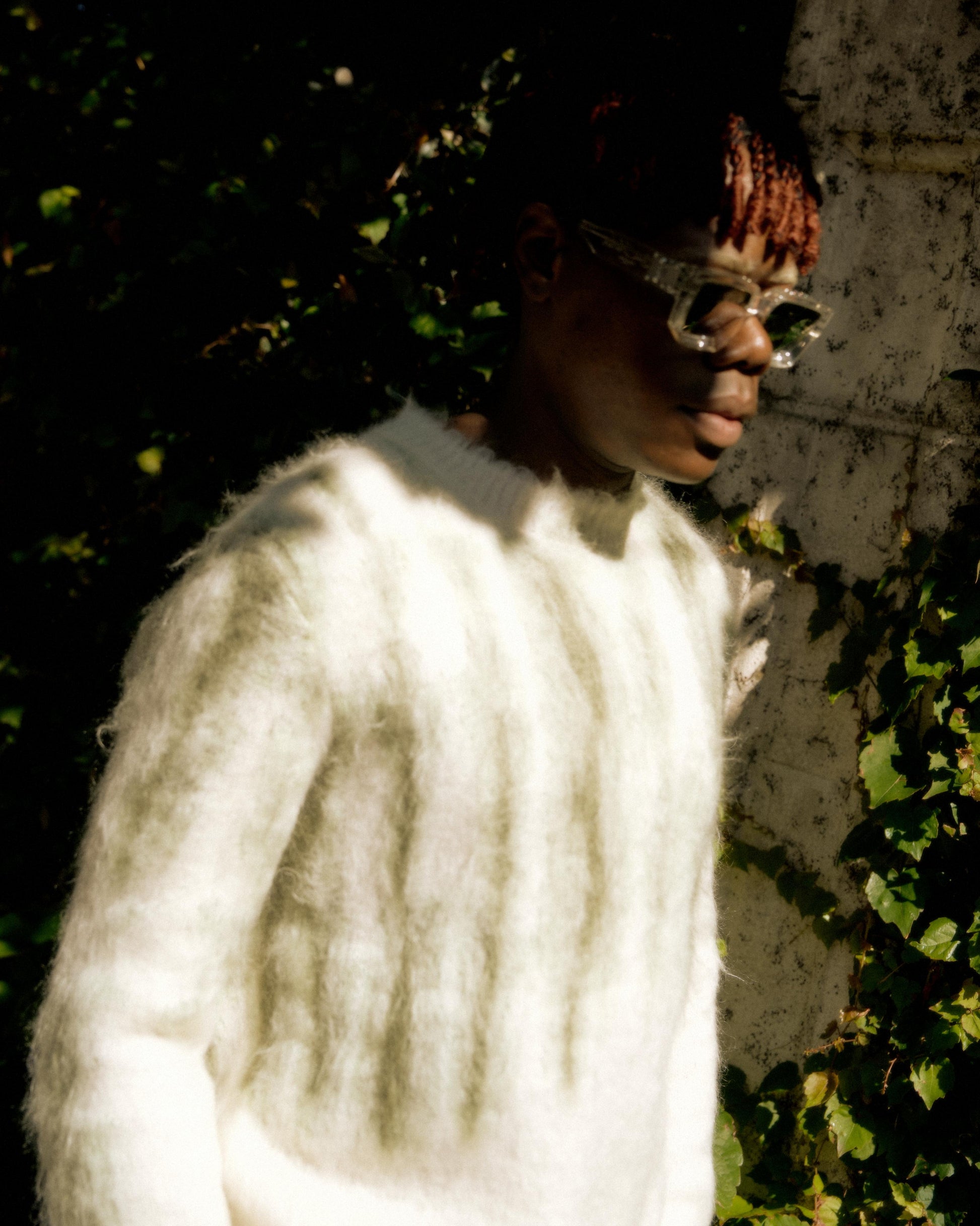 A person wearing a fuzzy white Louis Vuitton sweater and clear glasses is partially in shadow, standing against a vine-covered wall. 