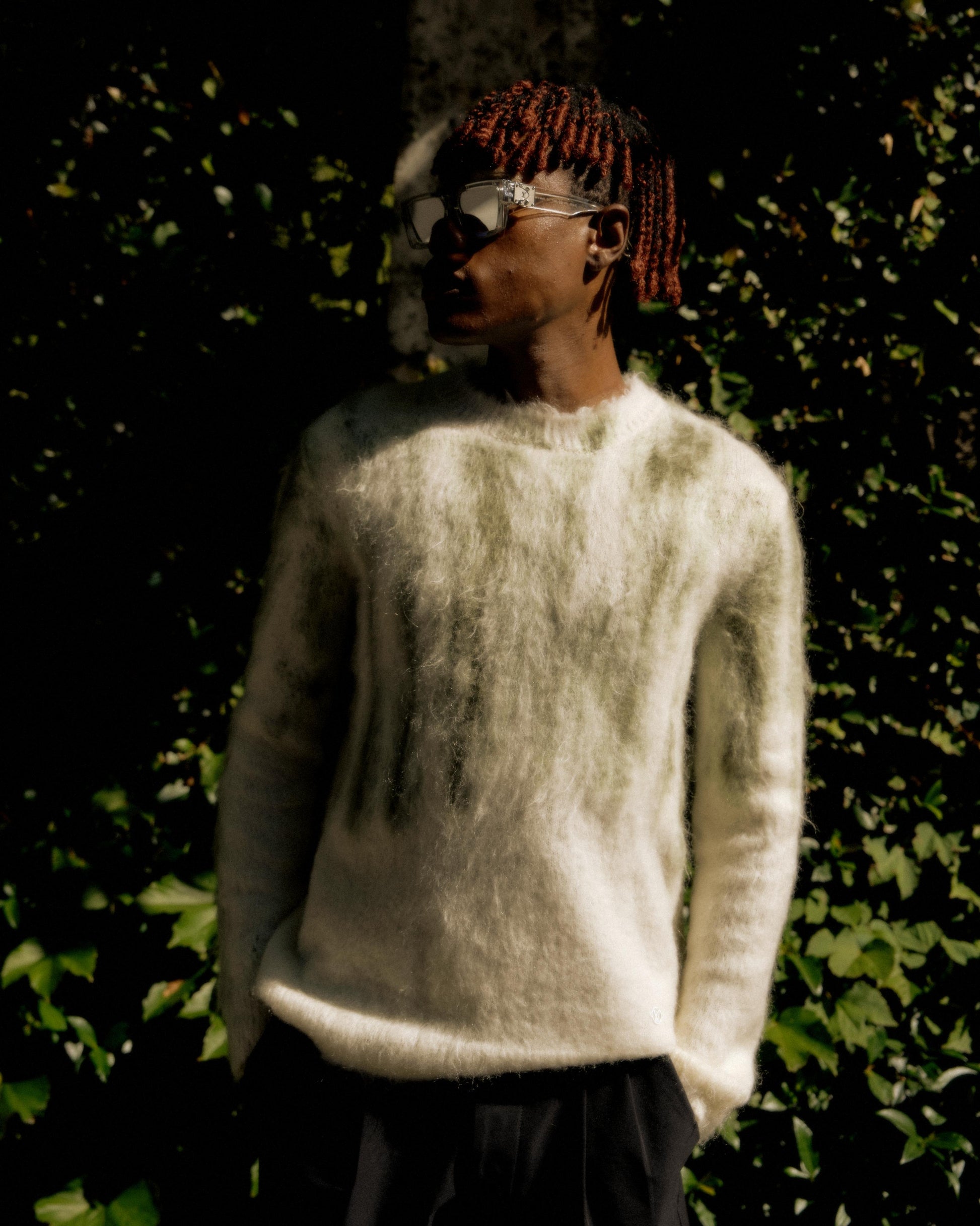 A person in a fuzzy white Louis Vuitton sweater and sunglasses stands against a background of lush green foliage. The lighting casts a dramatic shadow on their face.