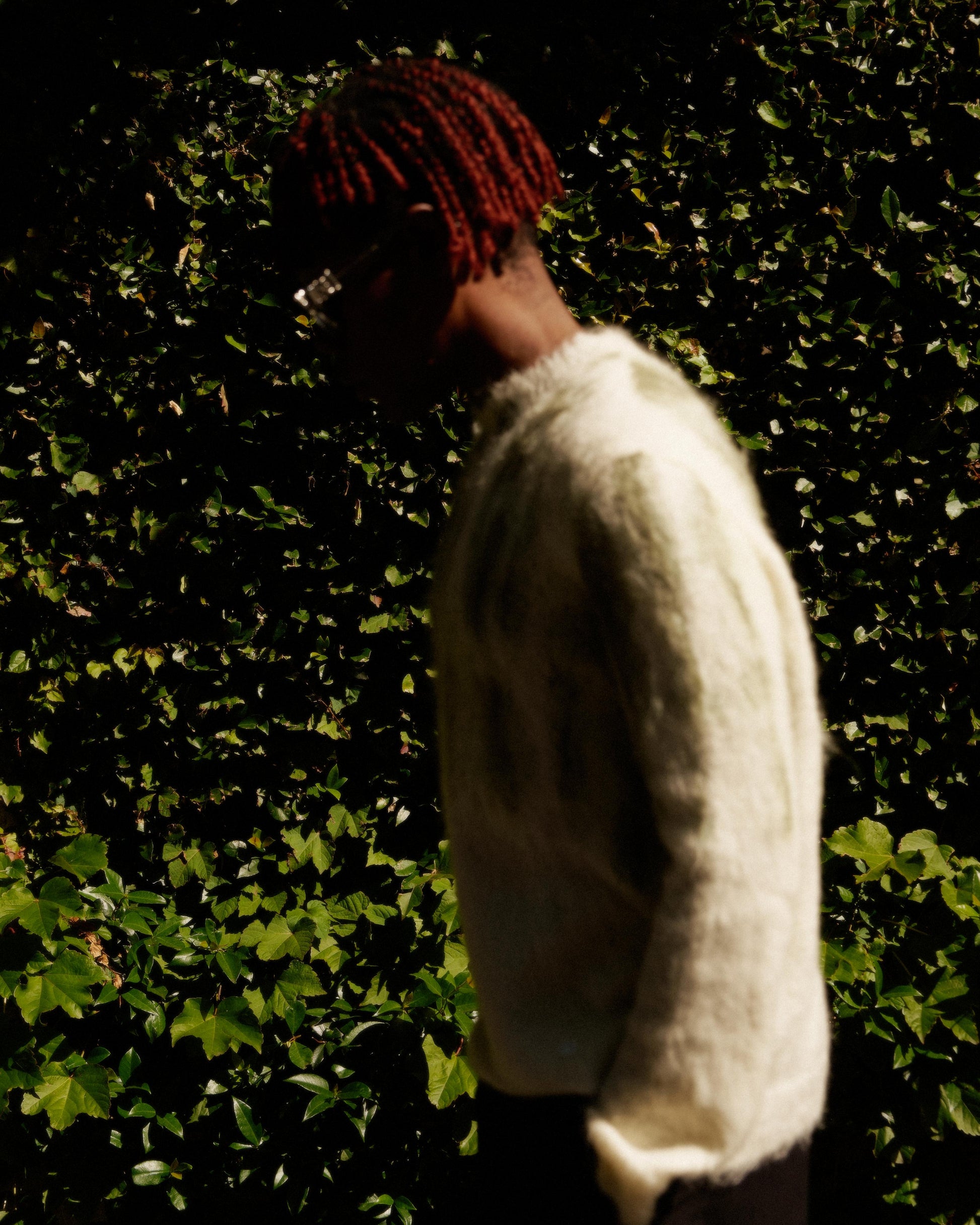 A person with red braided hair, wearing a fuzzy light Louis Vuitton sweater, stands in profile against a sunlit backdrop of dense green ivy, conveying a serene tone.