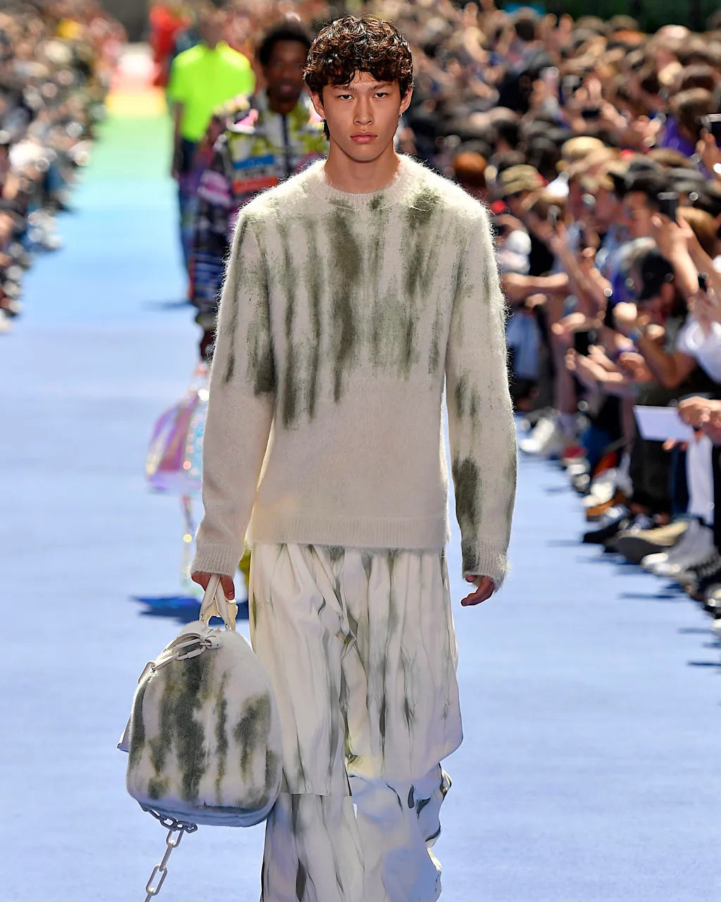 A model walks a vibrant Louis Vuitton runway, wearing a cream and green tie-dye sweater and pants, holding a matching bag. Crowd in background, sunny atmosphere.