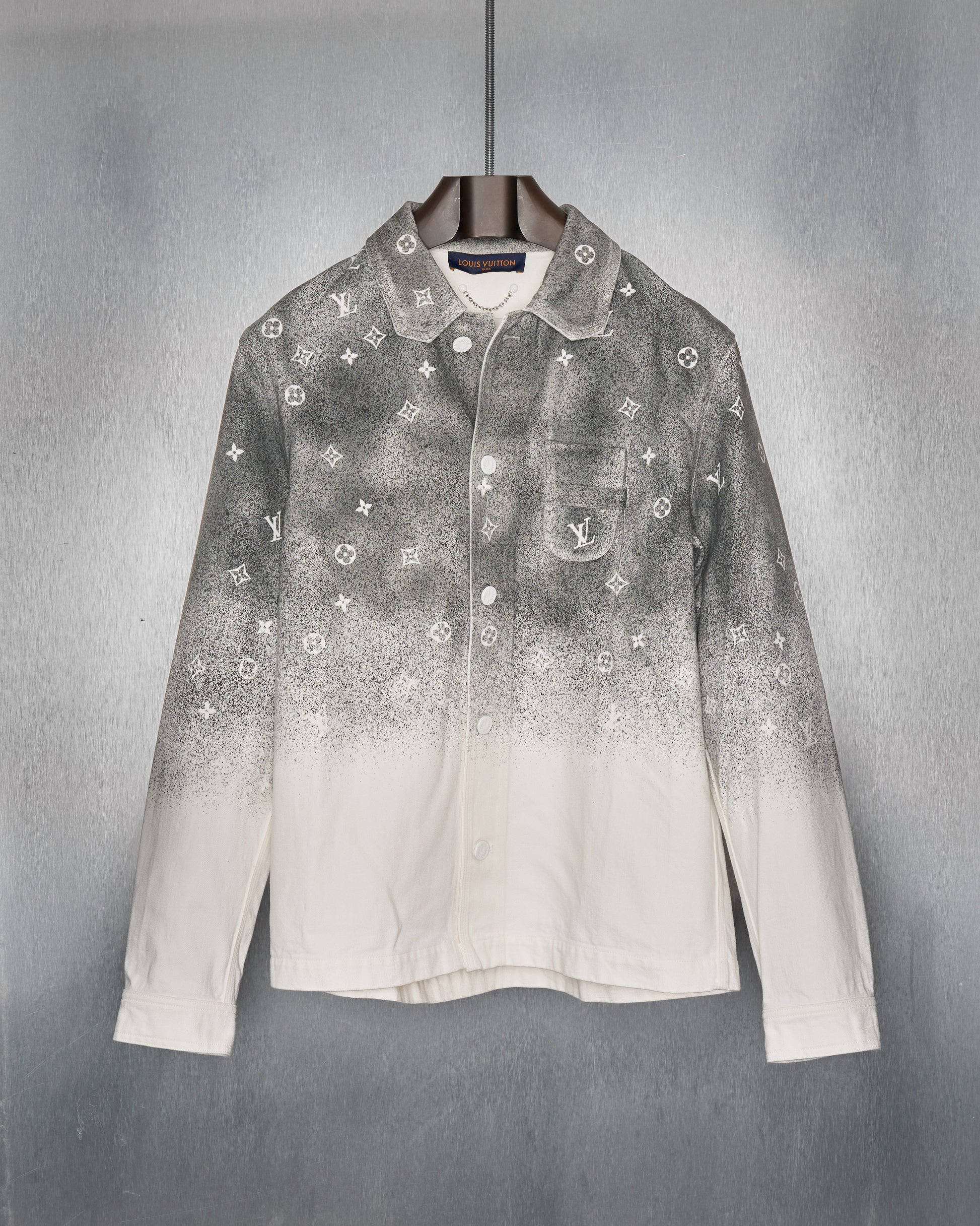 White Louis Vuitton jacket with a gradient black spray effect on top, featuring small, repeated white logos. It hangs on a wooden hanger against a steel backdrop.